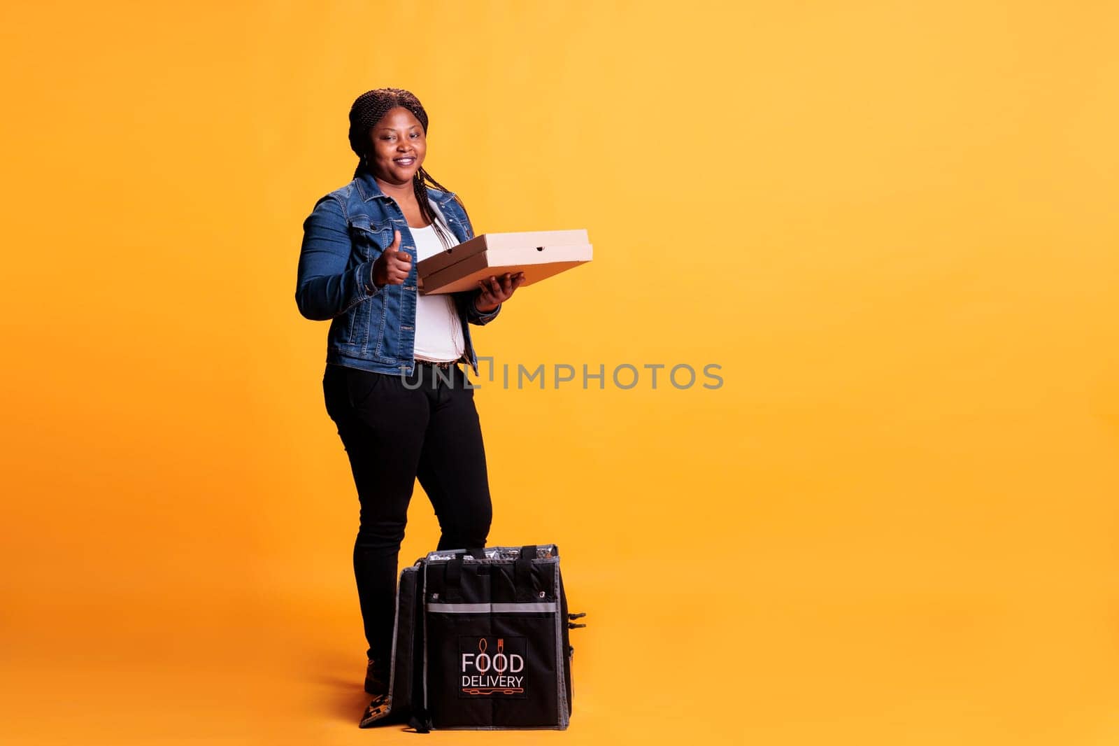 Confident deliverywoman holding pizza cartboard box while doing thumb up gesture at camera before delivery fast food order to client. Pizzeria courier carrying takeaway thermal backpack
