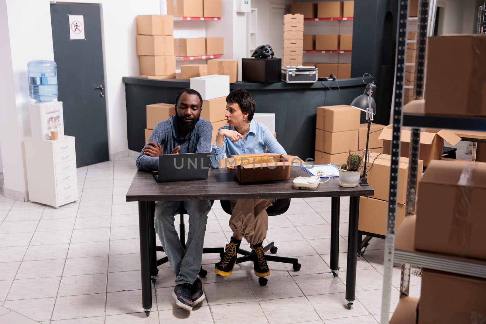 Workers sitting at table in storehouse checking transportation logistics on laptop computer before delivery trendy blouse. Fashion retail employees putting clothes in cardboard box