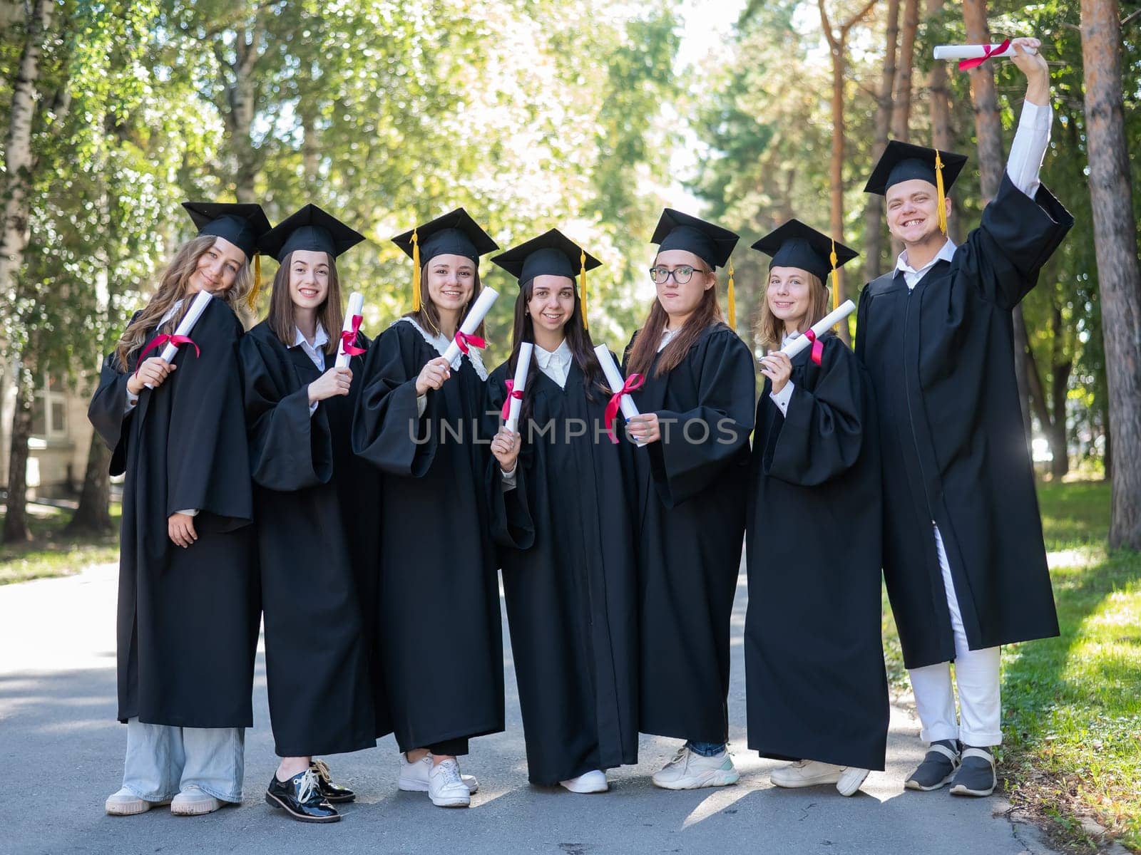 Row of young students in graduation gowns outdoors showing off their diplomas