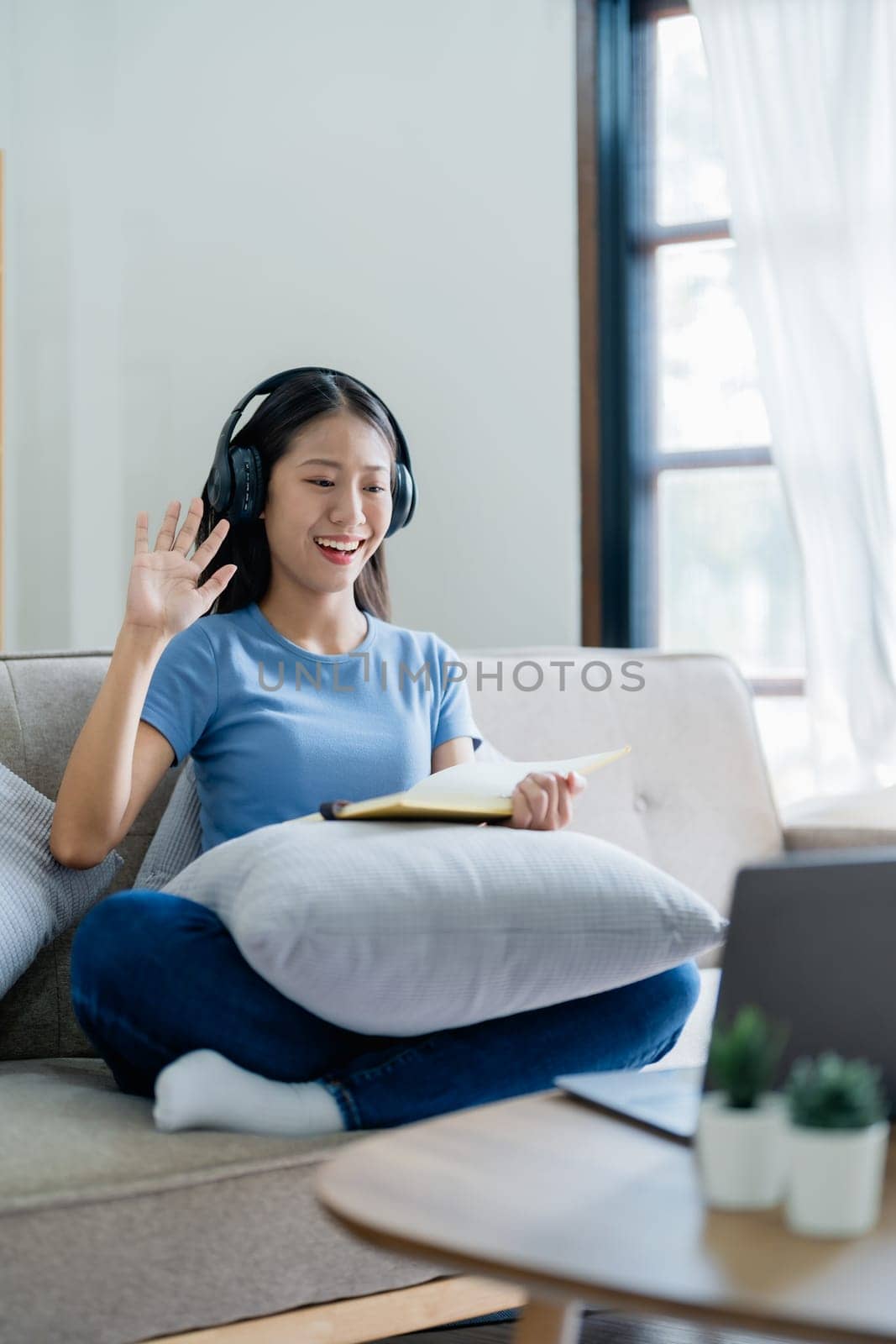 Asian young business woman is sitting on the sofa is wearing headphones video call conference with laptop computer raise hand saying hi team at home office, work from home. by Manastrong