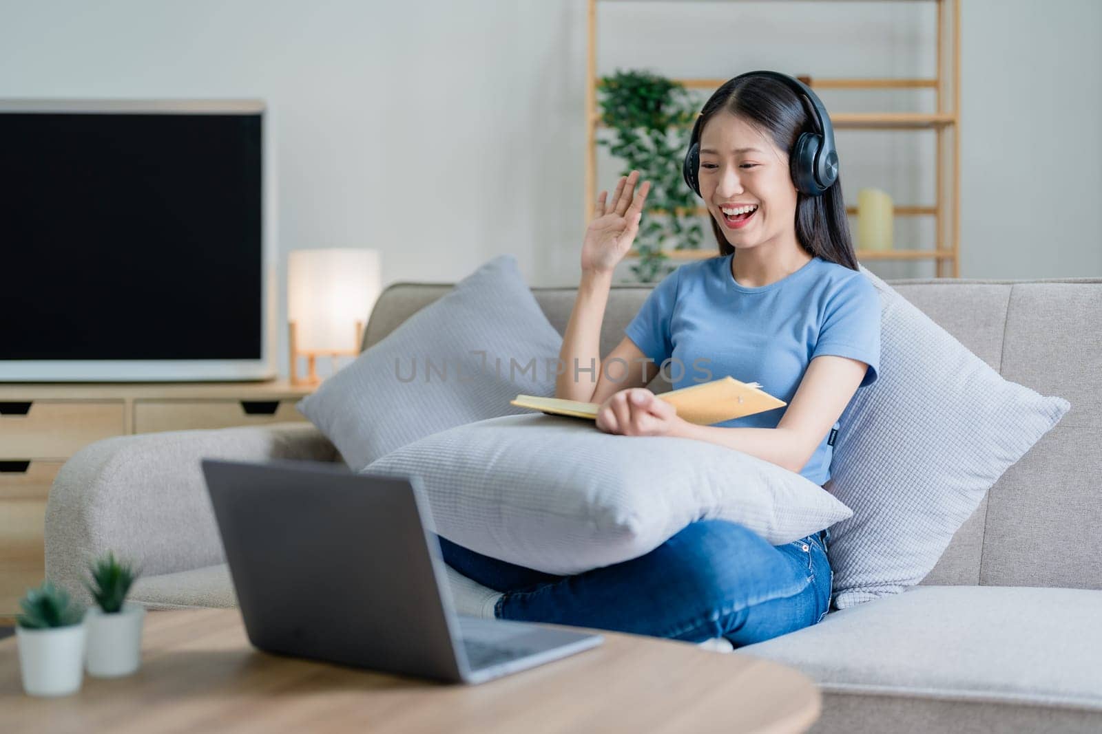 Asian young business woman is sitting on the sofa is wearing headphones video call conference with laptop computer raise hand saying hi team at home office, work from home