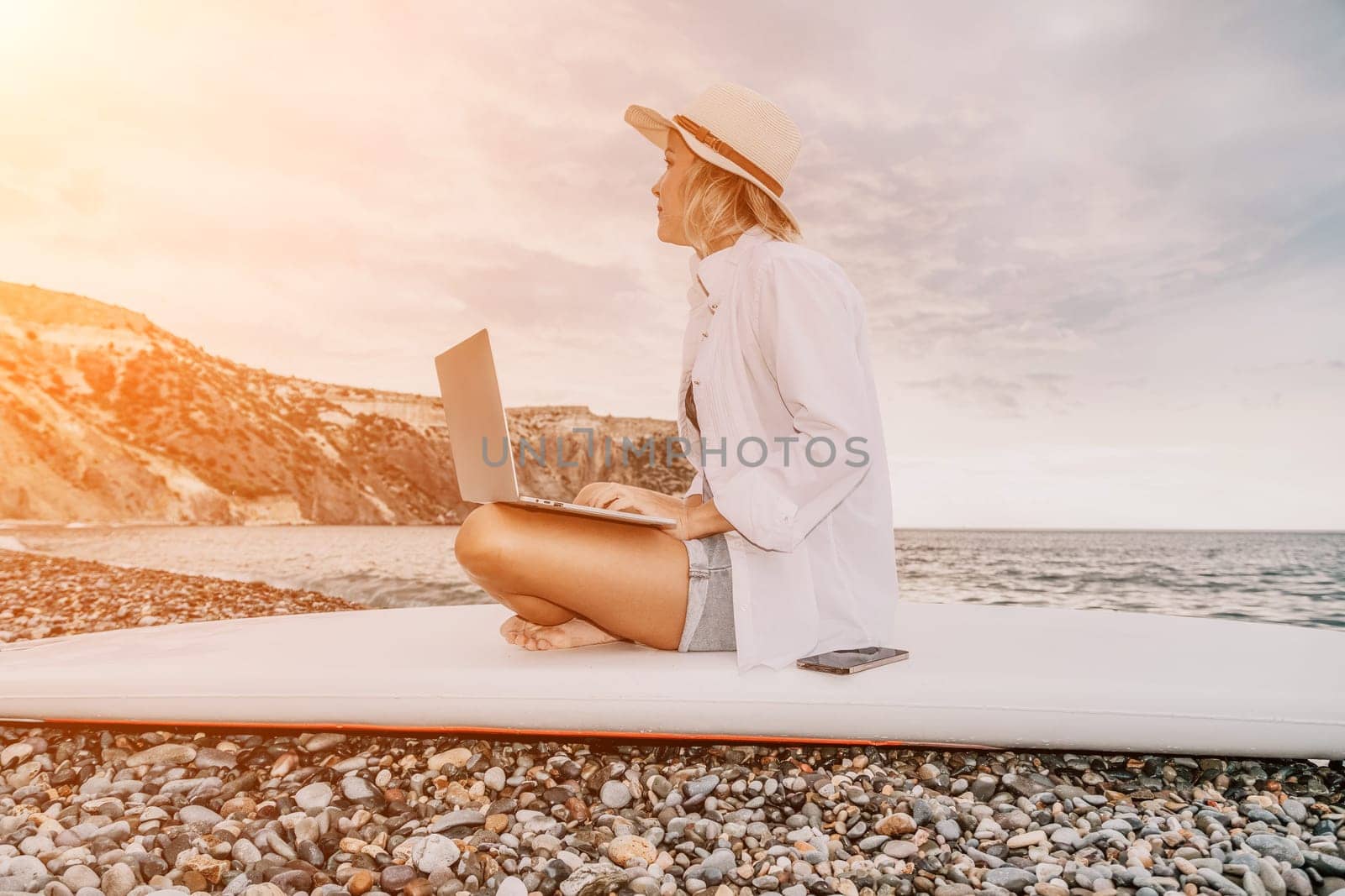 Digital nomad, Business woman working on laptop by the sea. Pretty lady typing on computer by the sea at sunset, makes a business transaction online from a distance. Freelance, remote work on vacation by panophotograph