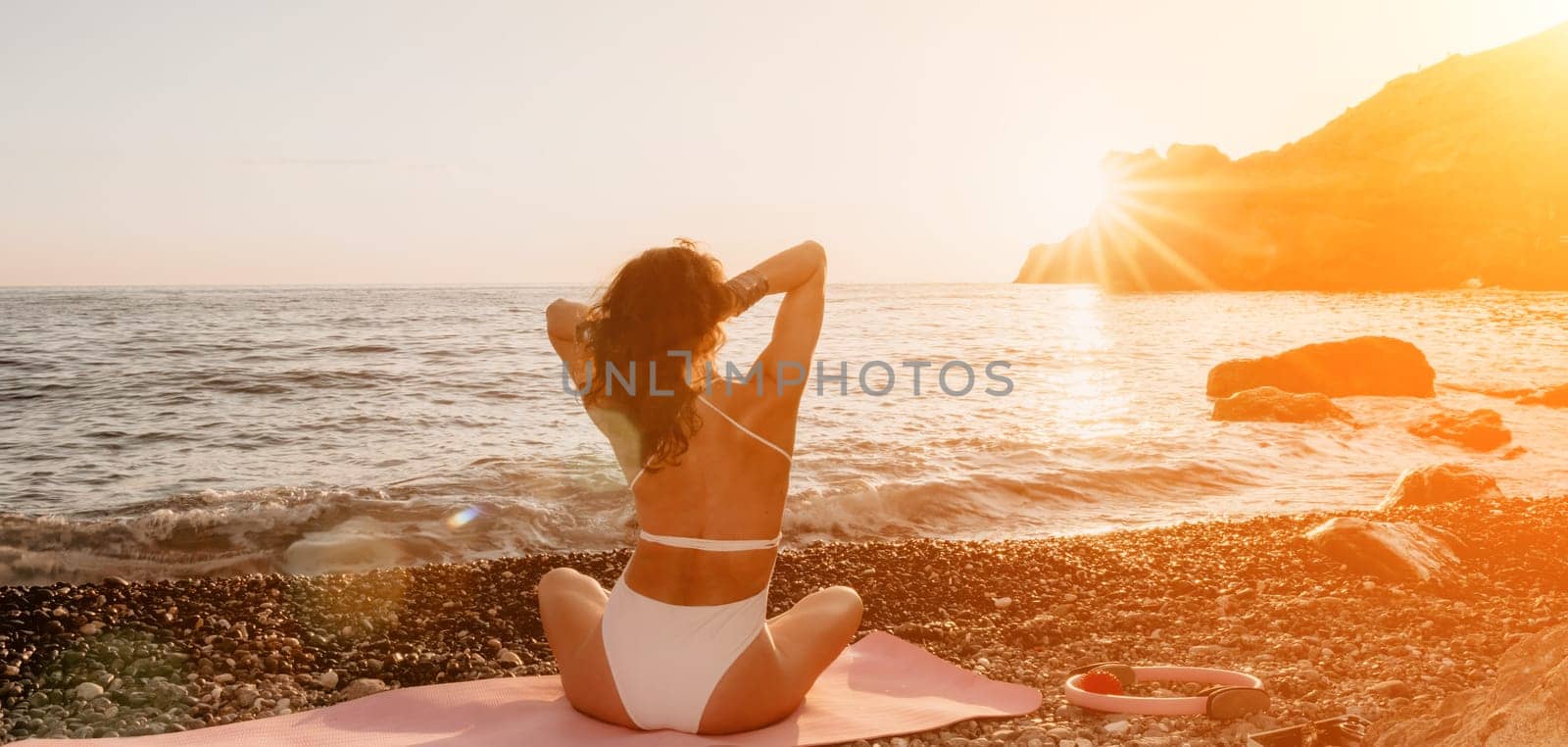 Young woman in swimsuit with long hair practicing stretching outdoors on yoga mat by the sea on a sunny day. Women's yoga fitness pilates routine. Healthy lifestyle, harmony and meditation concept.