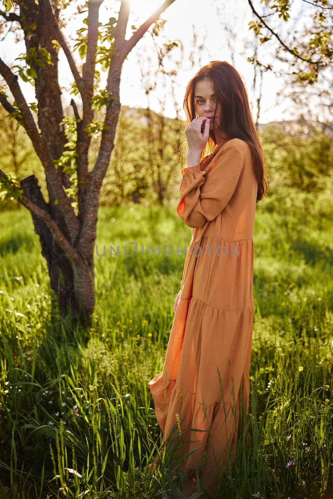 a sweet, thoughtful woman stands in nature near a tree in a long orange dress, illuminated from the back by the sunset rays of the sun and holding her hand near her face looks away. High quality photo