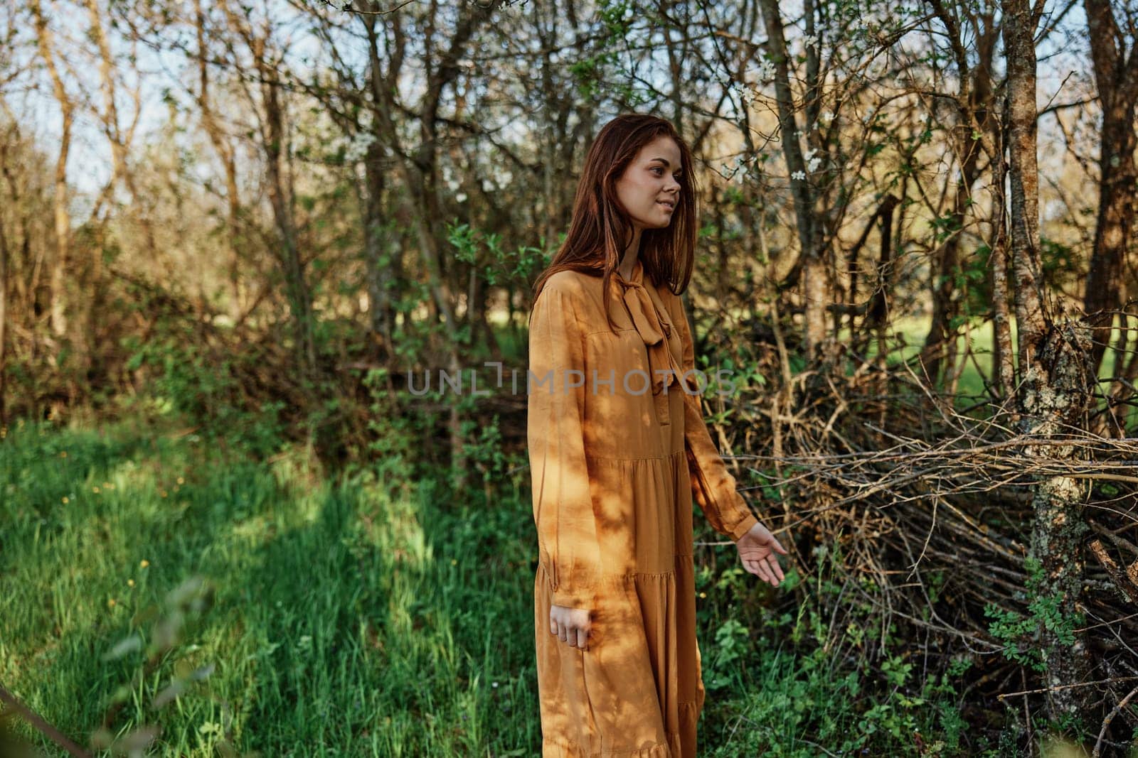 a beautiful, slender woman with long hair walks in the shade near the trees, dressed in a long orange dress, enjoying the weather and the weekend. High quality photo