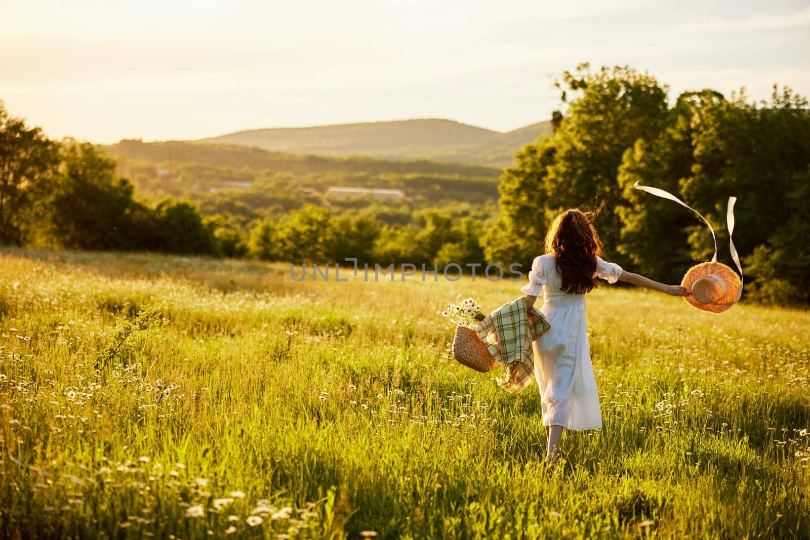 a woman in a light dress with a hat in her hands runs far into the field during sunset. High quality photo
