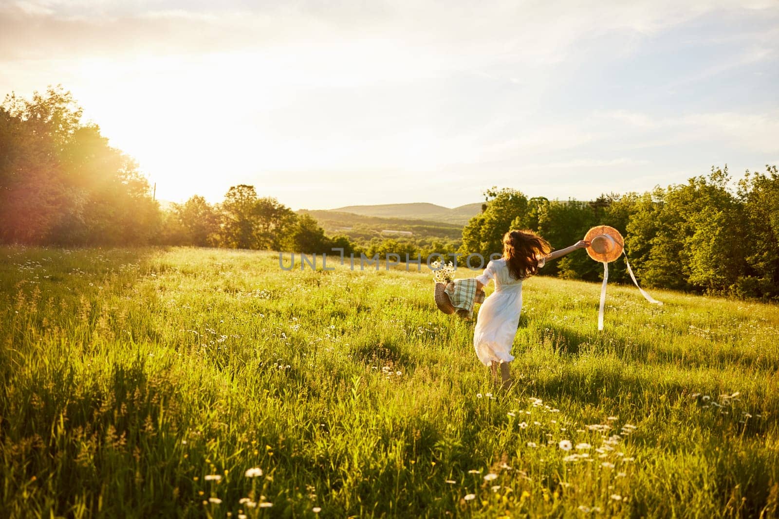 a woman in a light dress with a hat in her hands runs far into the field during sunset. High quality photo