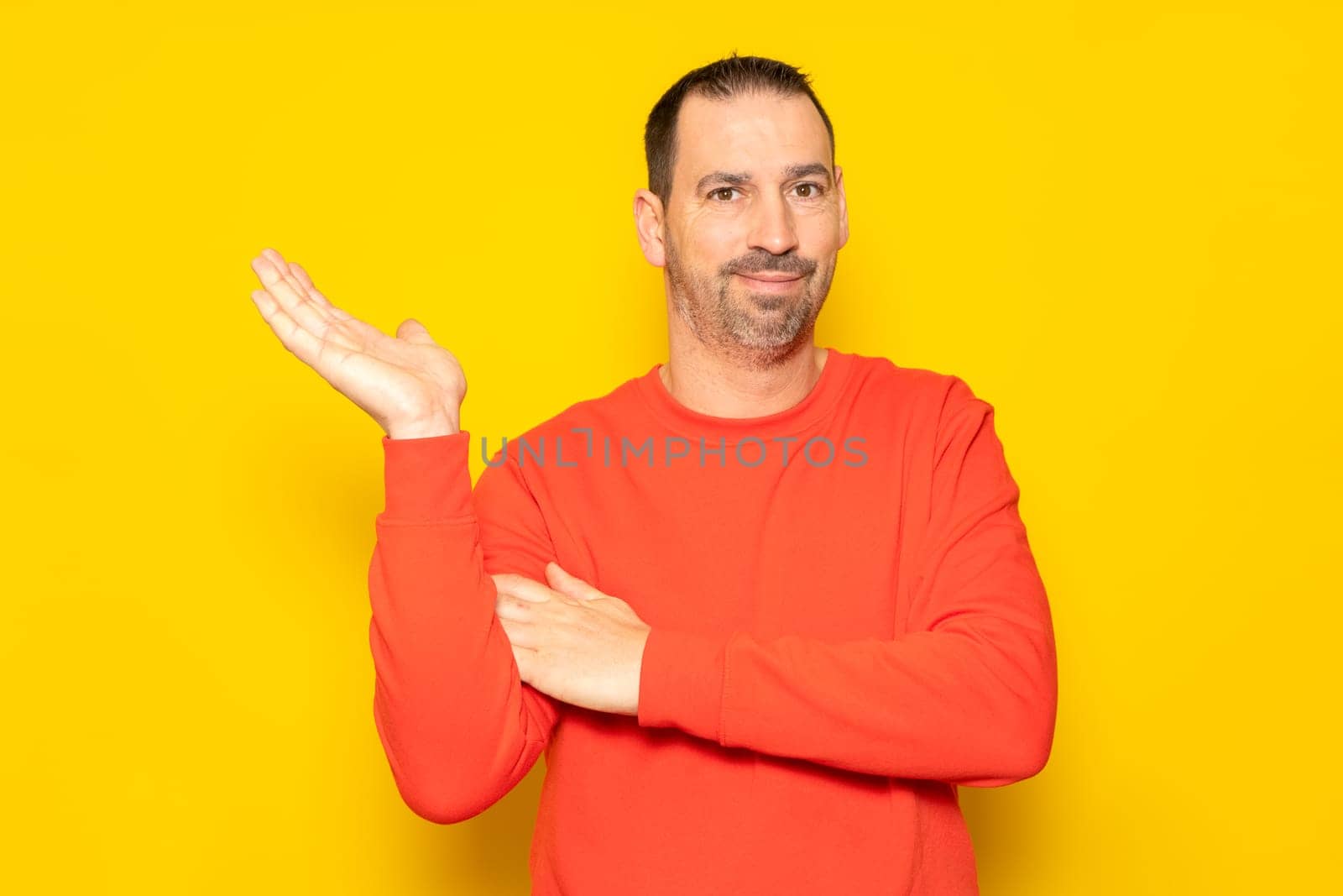 Hispanic man with a beard and red sweater in a friendly attitude offering something on the palm of his hand, isolated on a yellow background.