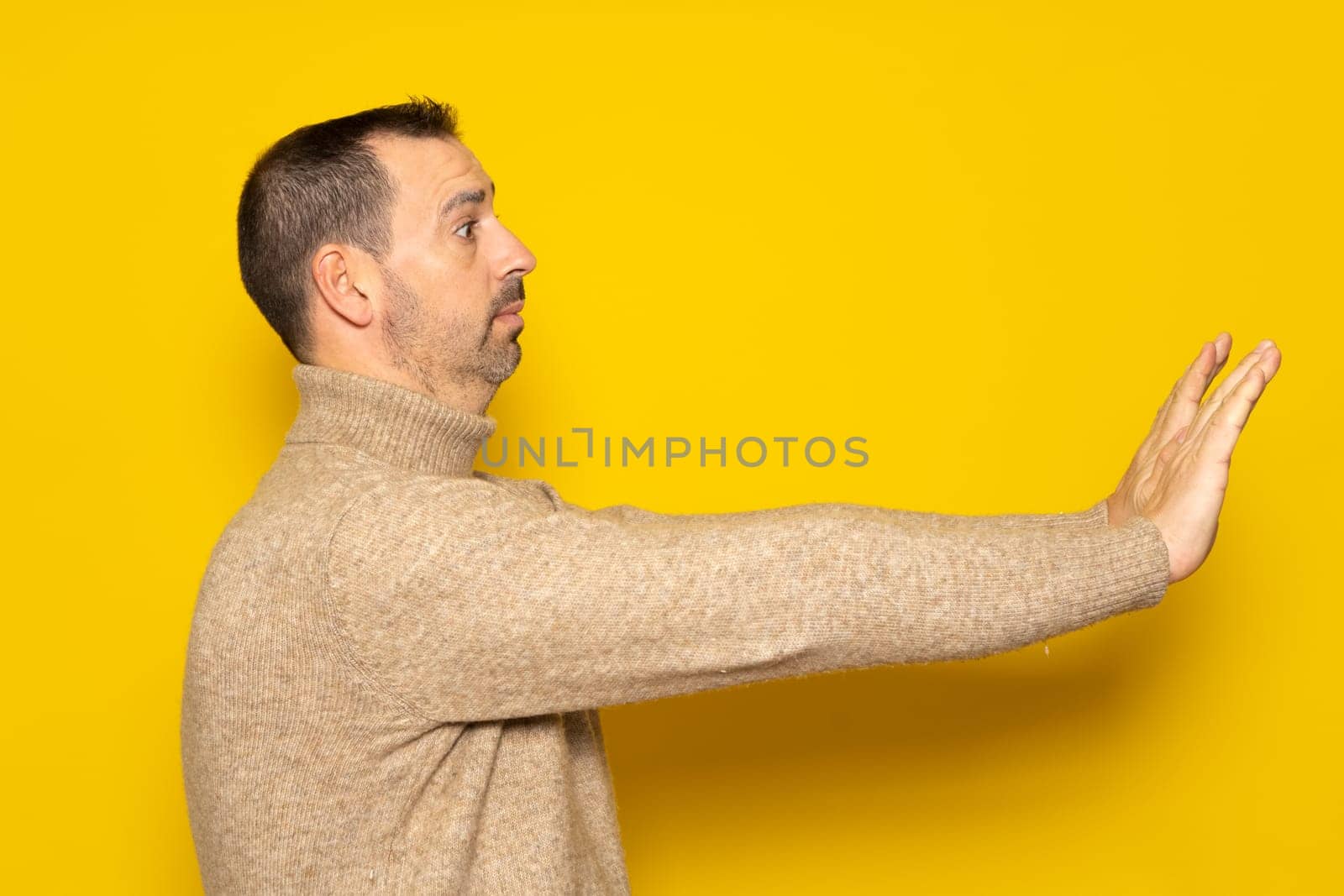 Side view of a scared man standing with a scared or worried face, looking at the camera and blocking with his hands. Indoor studio shot isolated on yellow background.