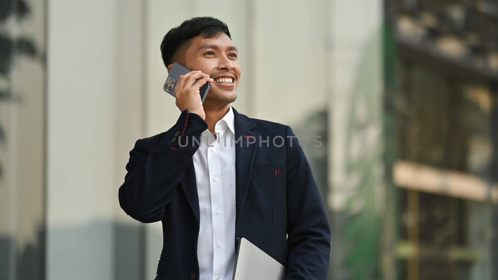 Pleasant millennial male entrepreneur in black suit having phone conversation while standing in front of business center.