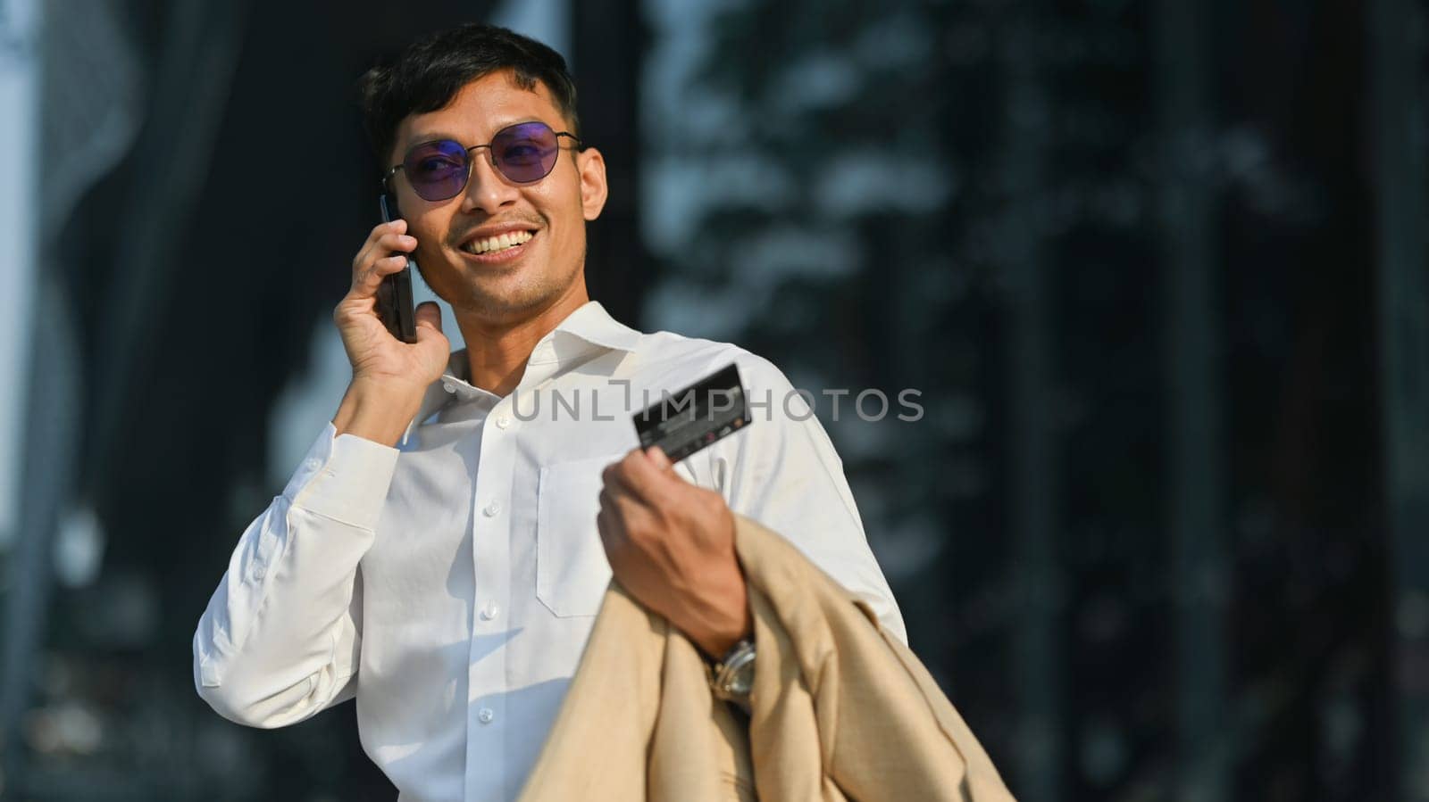 Smiling man holding credit card, calling to bank or customer service, while standing at outdoor in front of business center.