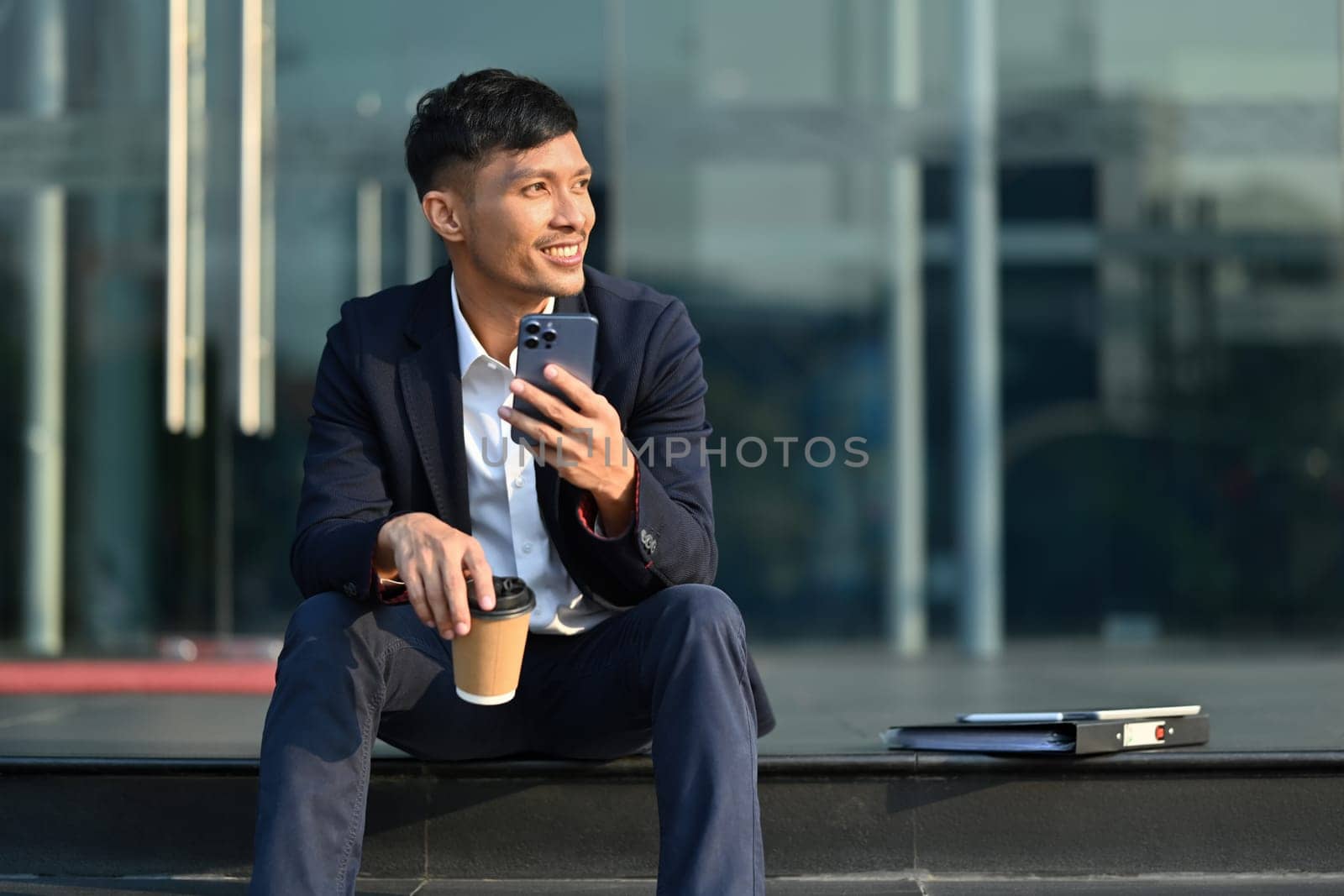 Attractive businessman in formalwear using smartphone in front of business center. Business, technology and lifestyle.