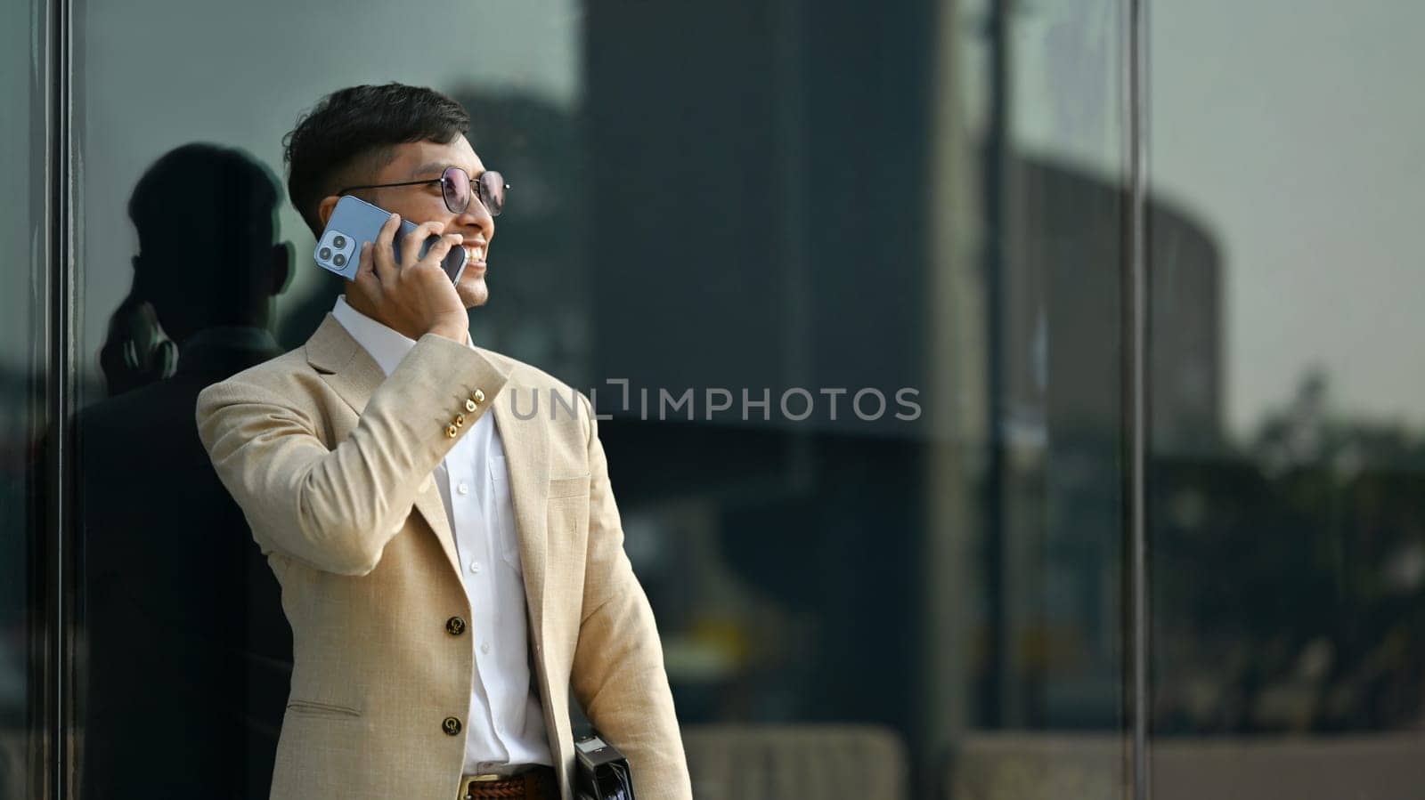 Portrait of adult man in elegant business suit having phone conversation while standing in front of modern office building.