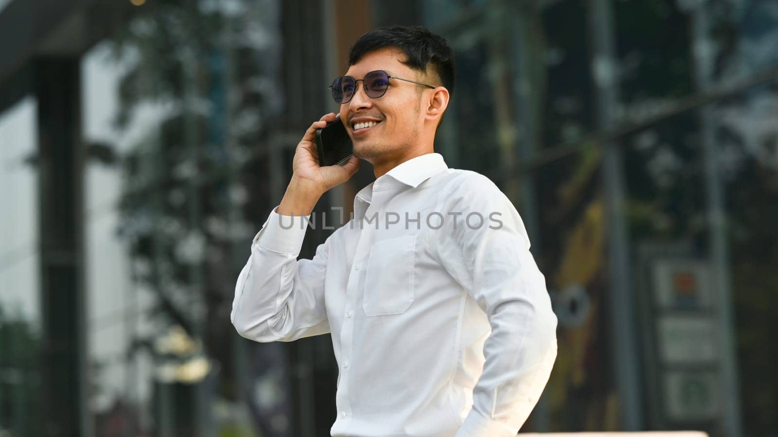Confident businessman in white shirt talking on mobile phone while standing outside of office in urban city.