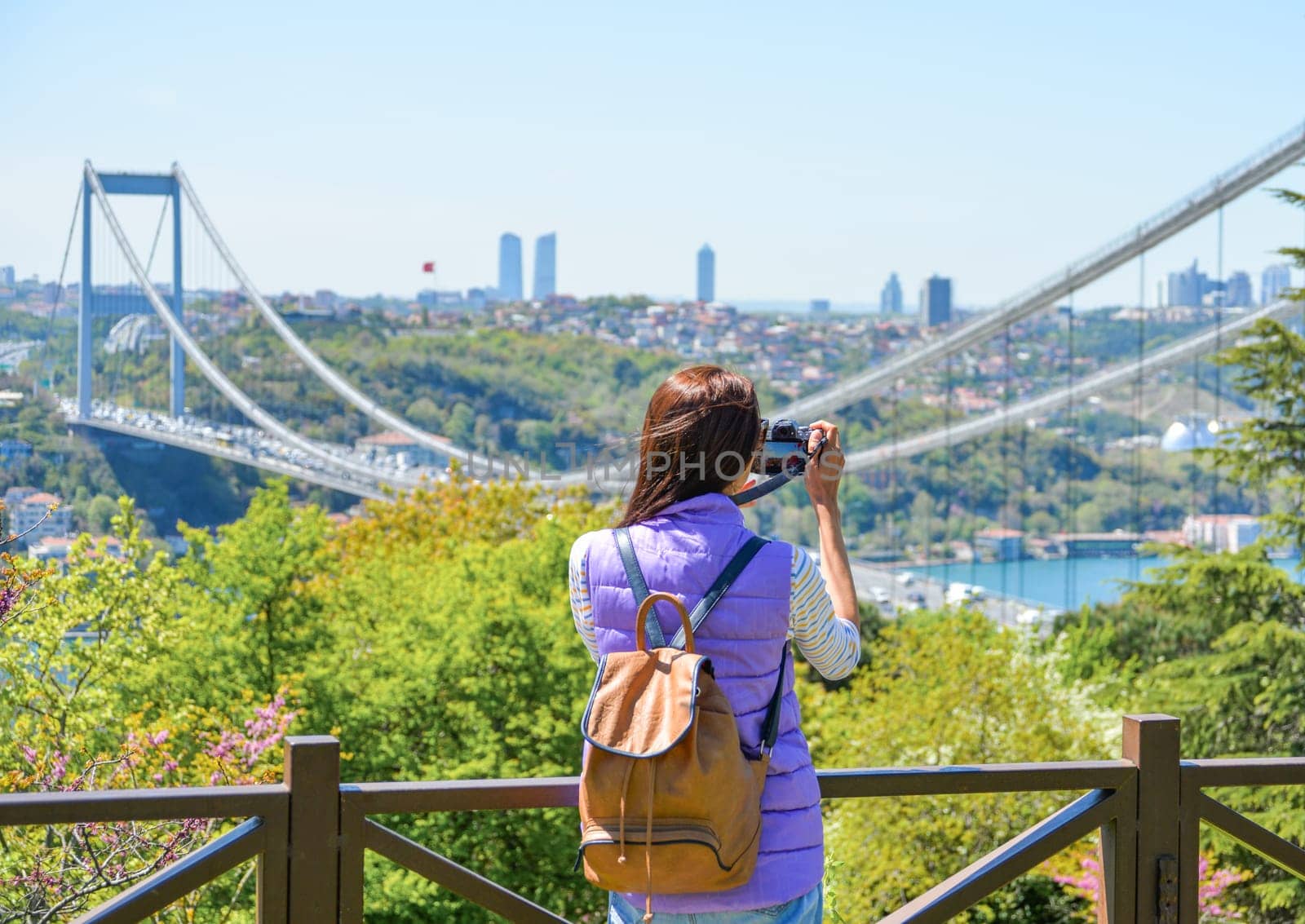 A young woman tourist with a backpack photographs the cityscape with a panoramic view of the Fatih Sultan Mehmet Bridge. Back view. Otagtepe Park.