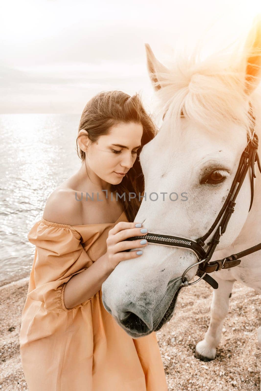 A woman in a dress stands next to a white horse on a beach, with the blue sky and sea in the background