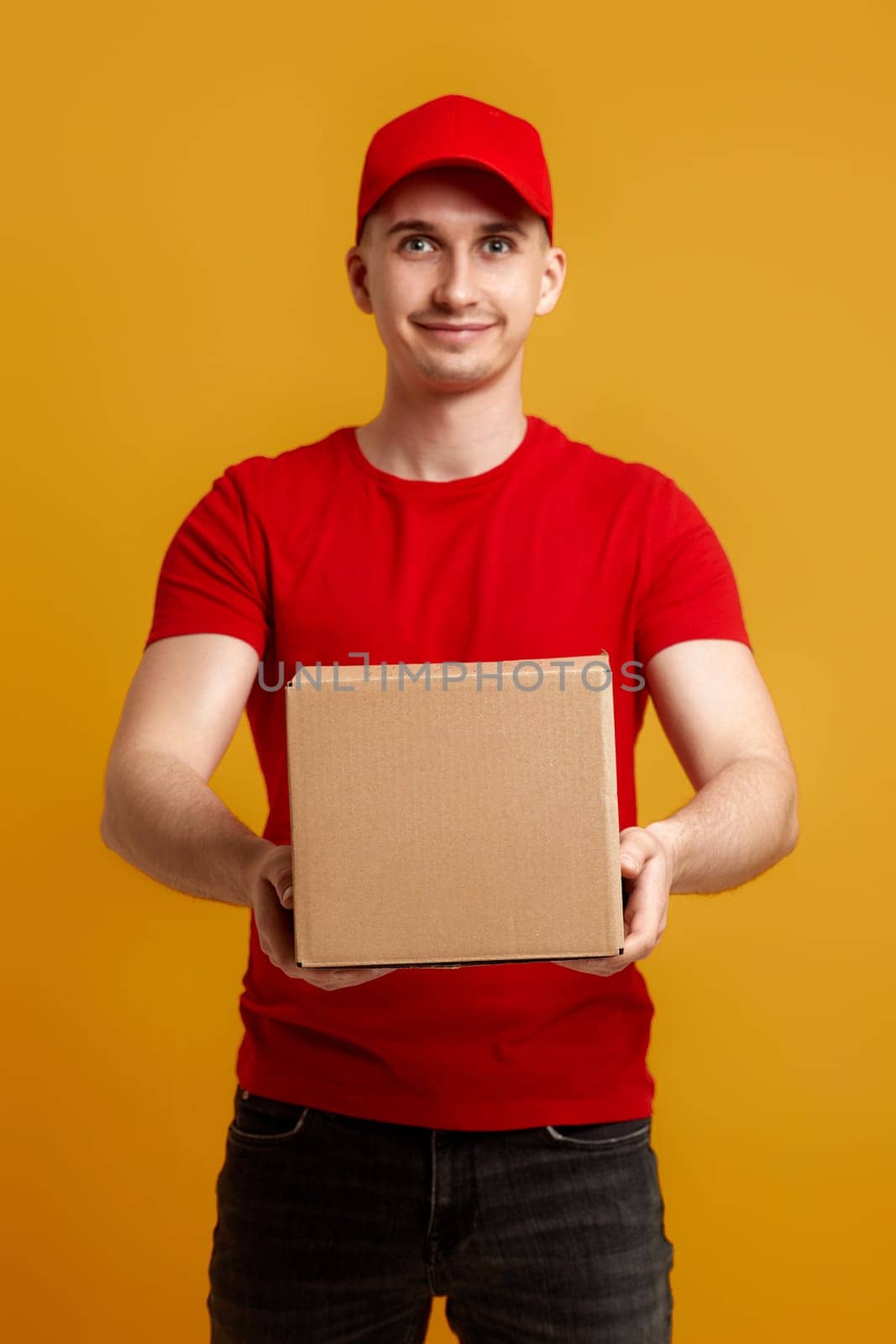 Delivery man employee in red cap, red t-shirt holding cardboard box on yellow background. focus on box