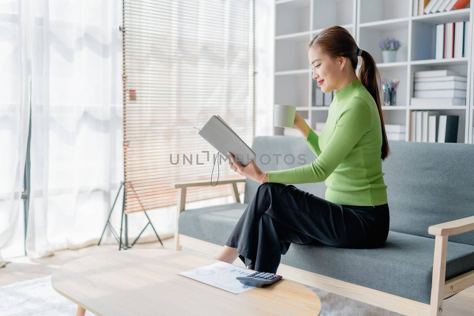 Beautiful young female for early in morning, writes notes in notepad, enjoys hot coffee from paper cup, sits on comfortable sofa, cozy domestic atmosphere. by Manastrong