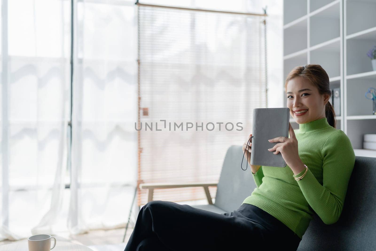 Beautiful young female for early in morning, writes notes in notepad, enjoys hot coffee from paper cup, sits on comfortable sofa, cozy domestic atmosphere. by Manastrong