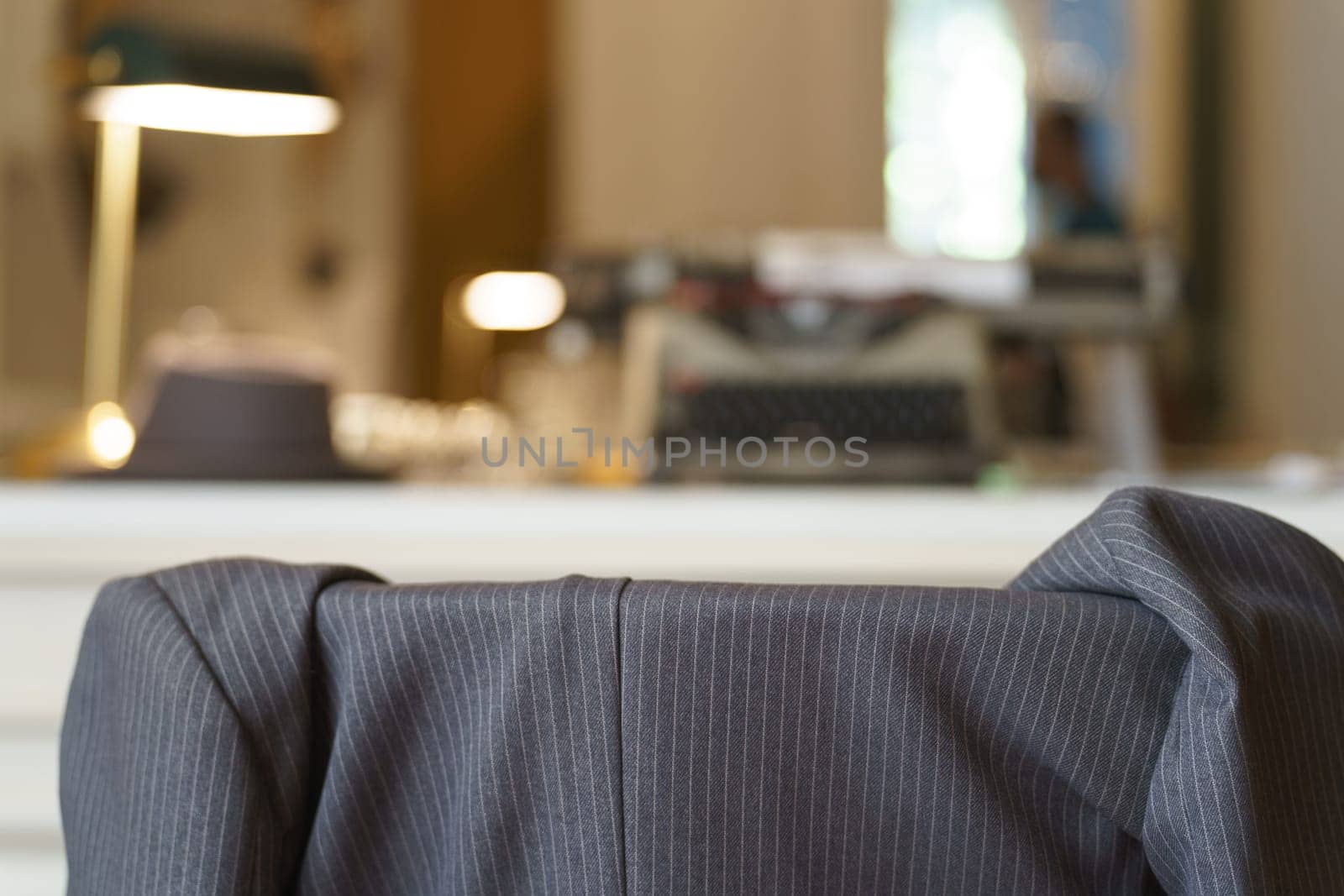 Antique writer's workspace with a typewriter, an empty chair with a stylish jacket hanging on it, and a blurred background of a hat, lamp, and typewriter. Perfect for adding text to create a lost writer's story.