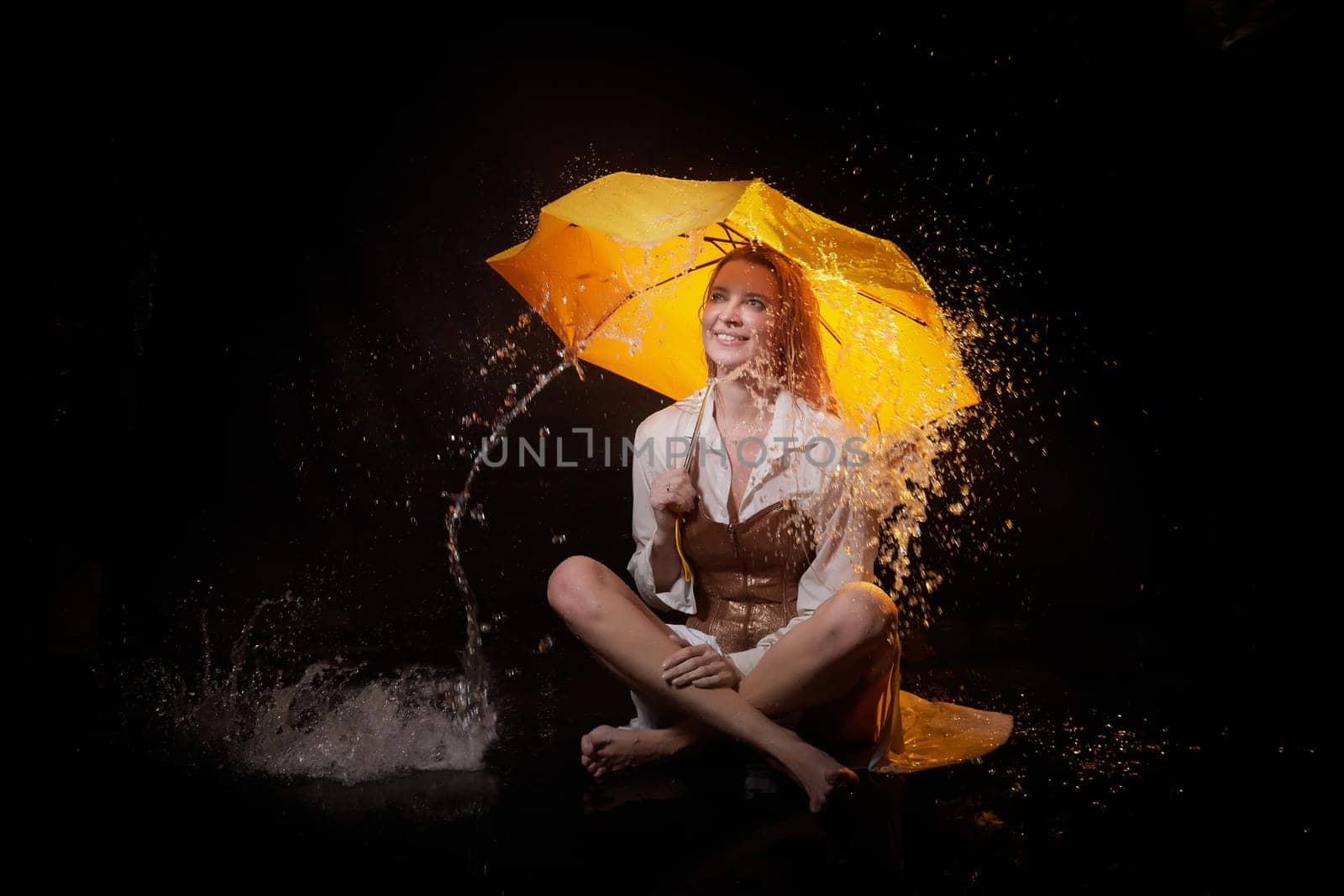 Beautiful sexy skinny girl in a dark hall with splashes of water and yellow umbrella under rain. Female Model blonde woman posing with water on black background in the studio