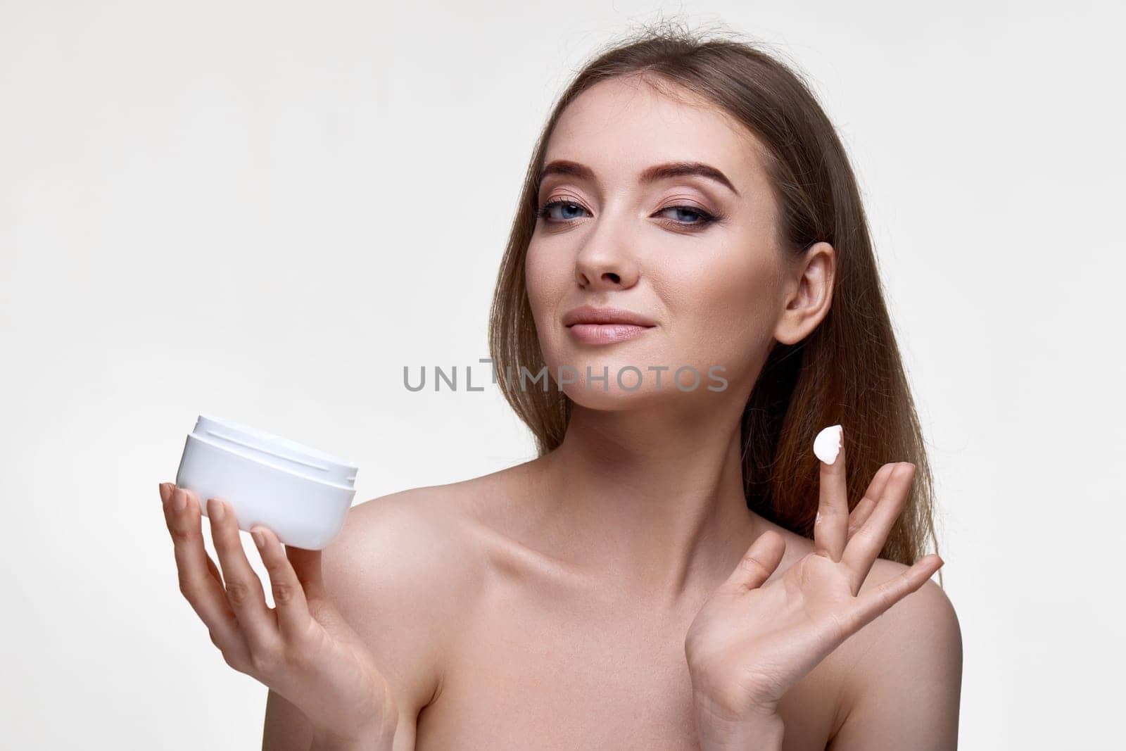 beautiful smiling girl applying skin care cream on her face and holding jar of cosmetic product on white background