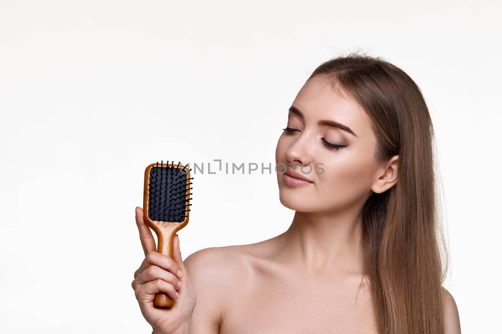 Beautiful brunette woman showing wooden brush and recommend haircare product for healthy long hair isolated on white background