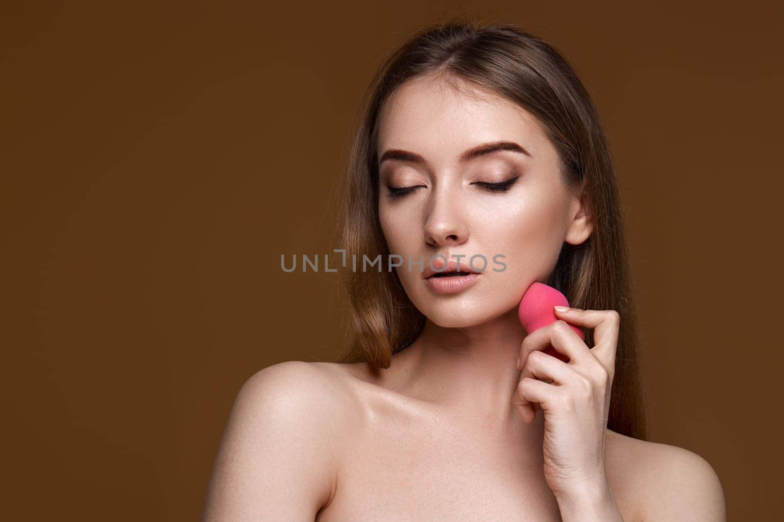 smiling young woman applying foundation on skin with beauty blender isolated on beige background
