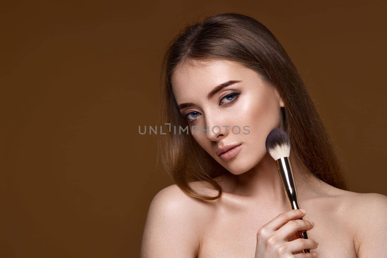 young woman applying blush on cheeks with make-up brush on beige background. copy space