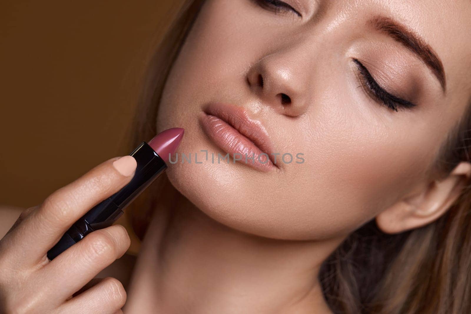 beautiful young smiling woman holding nude pink lipstick on beige background