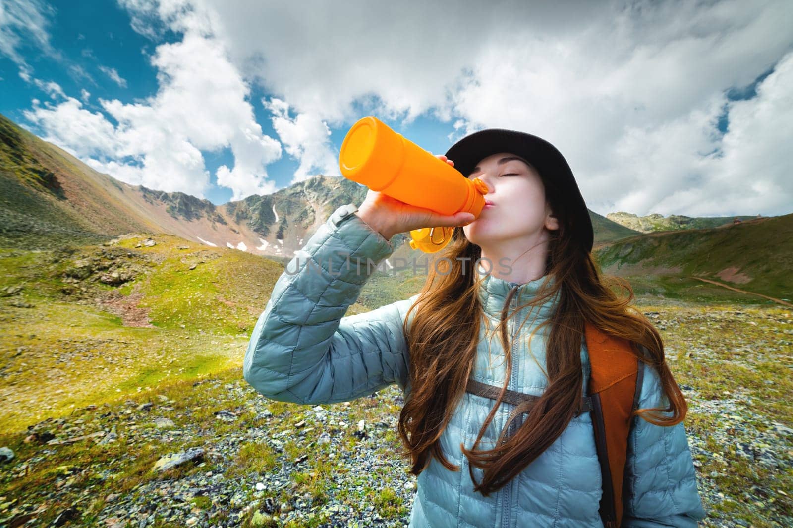 A woman takes a break to drink from a water bottle while hiking. Young hiker drinks water in a mountain valley.