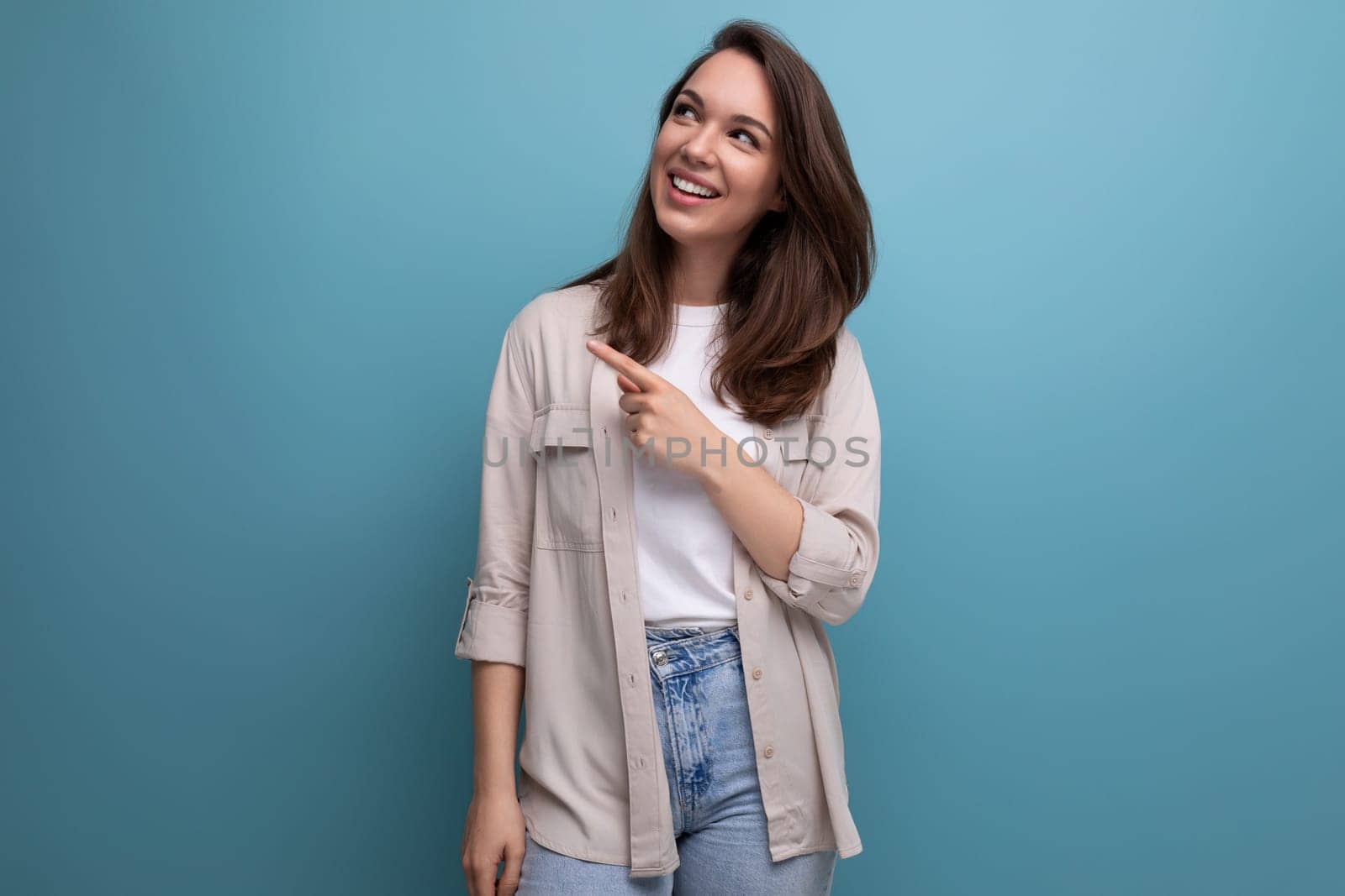 charming brunette 30s woman in shirt and jeans smiling on blue background.