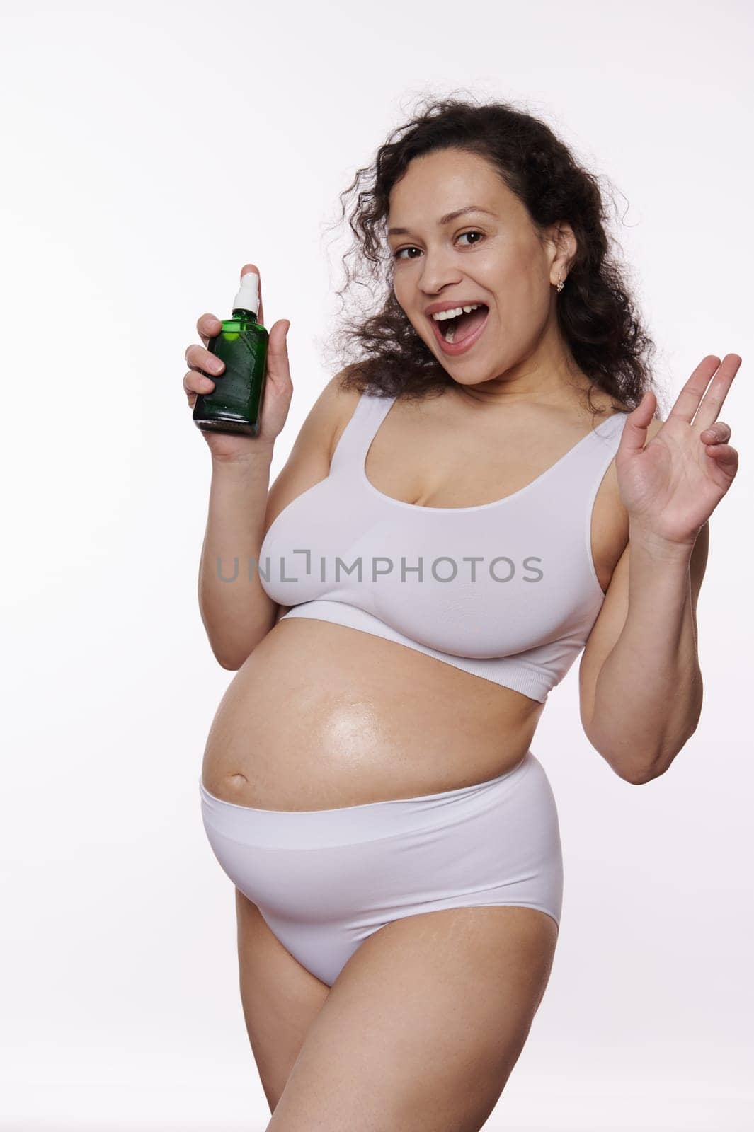 Smiling pregnant woman in white underwear, holding a bottle with cosmetic product, applying massage oil on belly against isolated white background. Preventing stretch marks. Skin Care during pregnancy