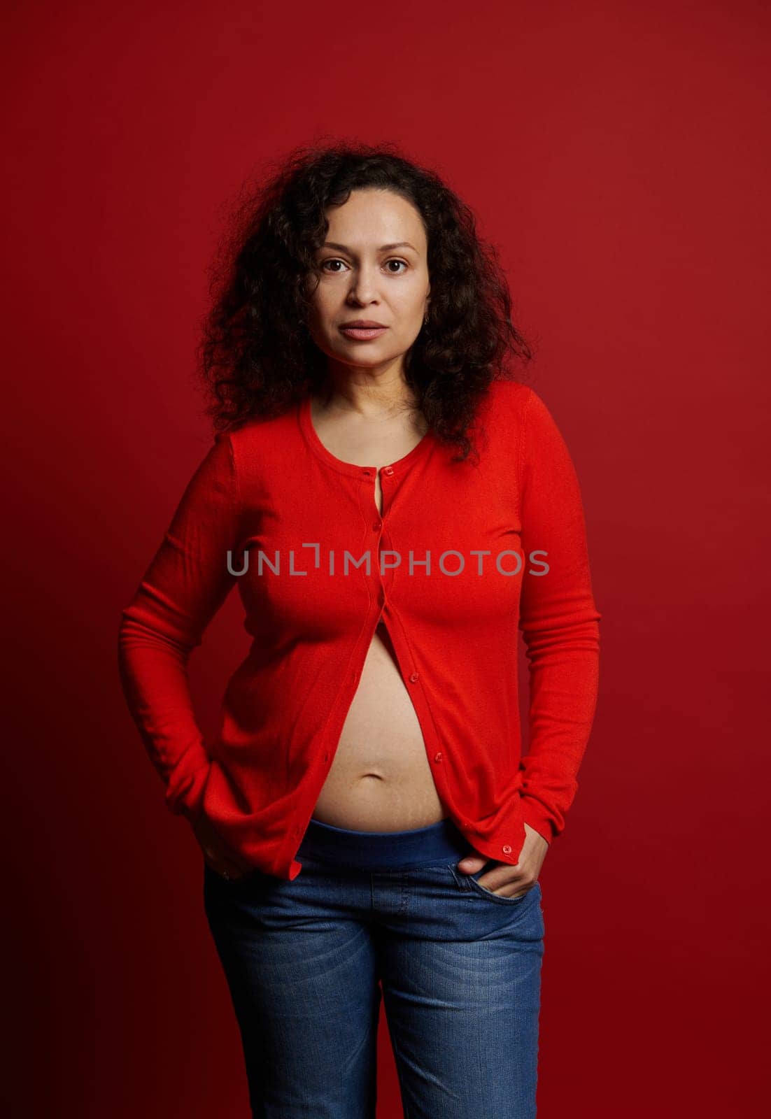 Confident stylish Latin American young pregnant woman wearing unbuttoned red shirt and blue jeans, posing bare belly on red backdrop, looking at camera. Expectant mother. Pregnancy fashion. Maternity