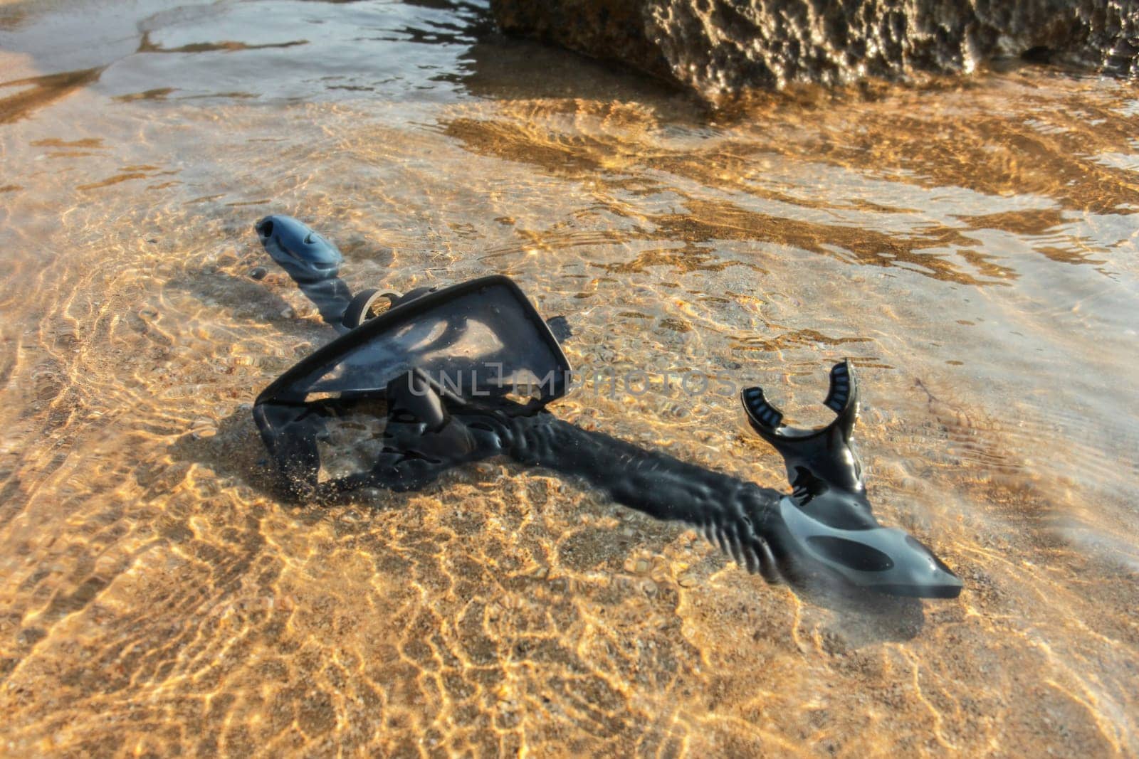 Black snorkelling tube and diving mask / goggles on sand in shallow sea water.