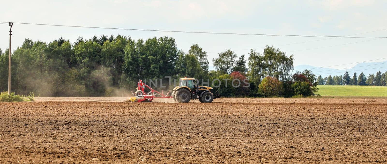 Tractor sowing on empty field, small trees in background. Wide agriculture banner. by Ivanko