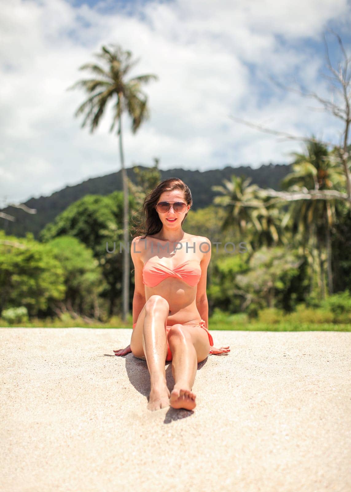 Young woman in bikini and sunglasses sitting on beach sand, looking into camera, palms and sky with clouds behind her.