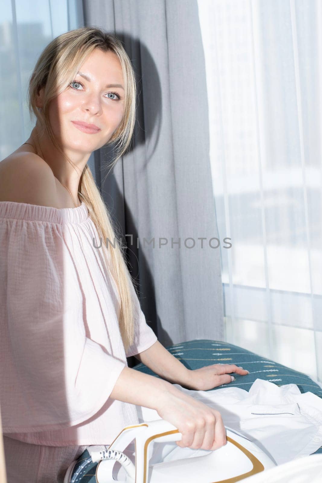 People. A beautiful Caucasian girl with long blond hair irons clothes on an ironing board against the background of a window, in pink home clothes with a bare shoulder. Concept.