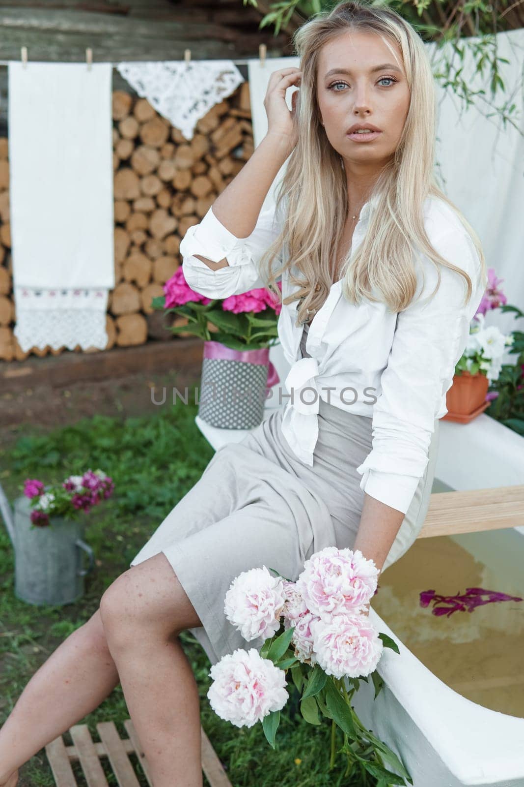 A woman is sitting on a cast-iron bathtub in the courtyard of a country house next to a bush of flowering peonies. The concept of summer, country life, a bathroom on the street in a blooming garden in the country