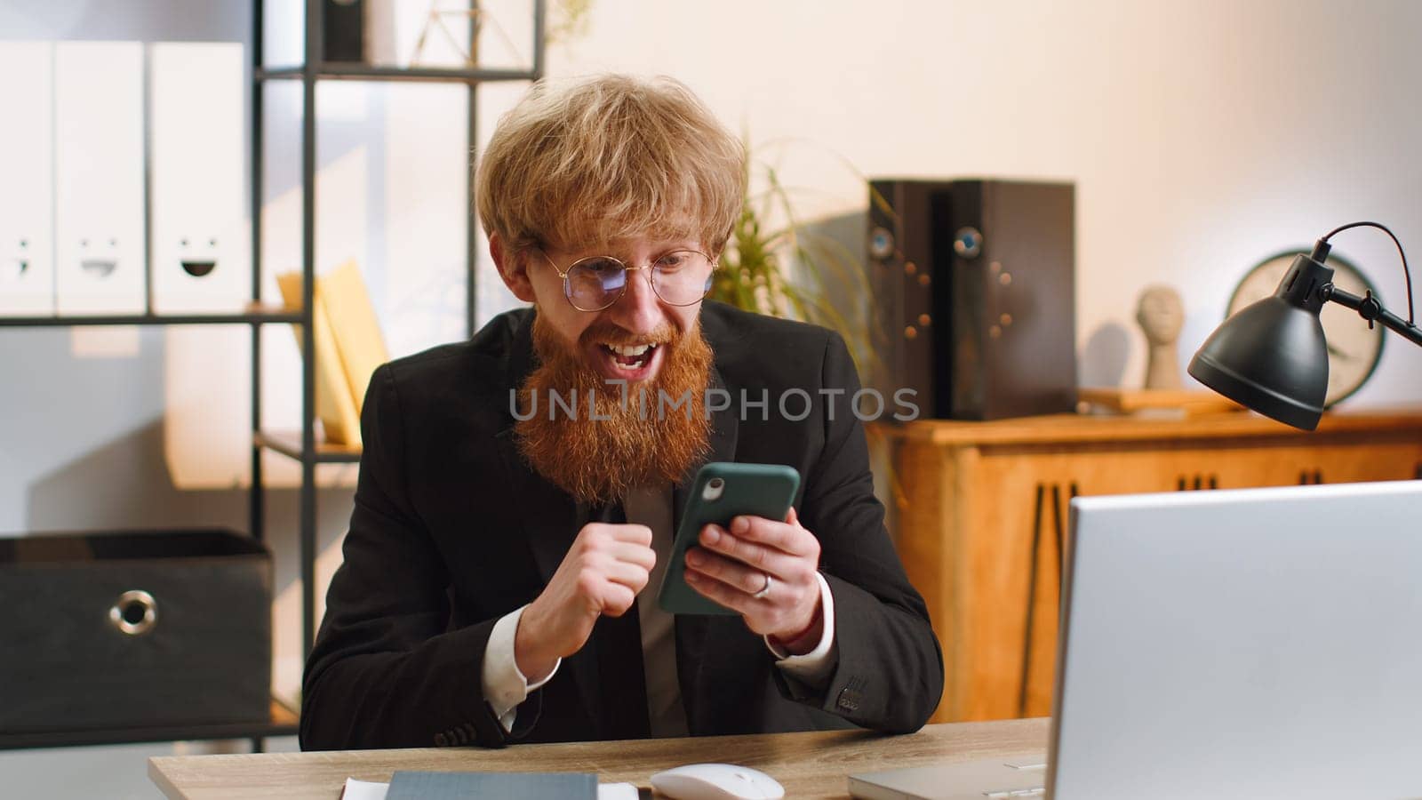 Oh my God Wow. Happy amazed young businessman guy working on smartphone shocked by sudden victory game winning lottery jackpot achievemen good news. Bearded redhead man freelancer at home office