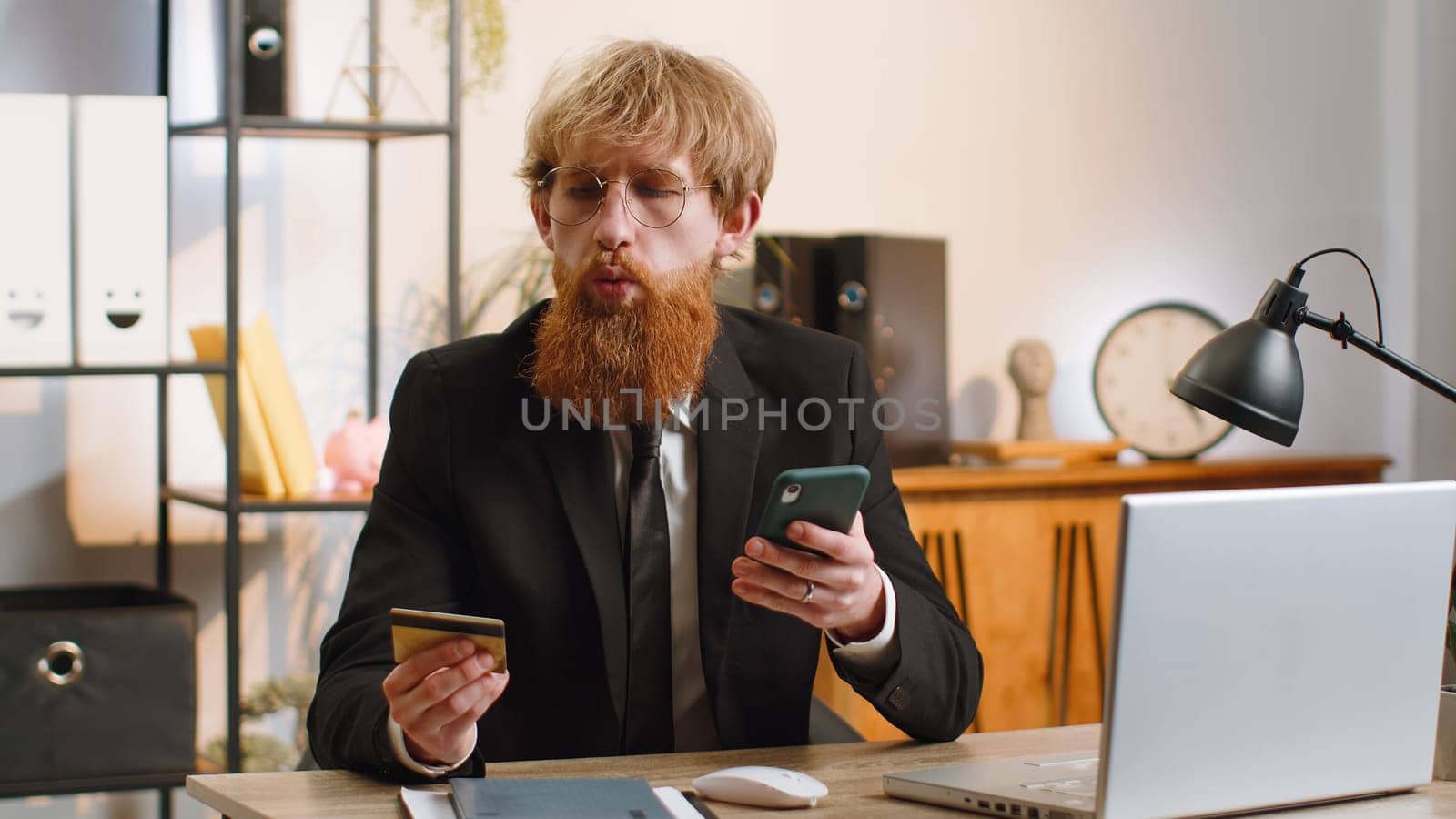 Bearded businessman programmer software developer working making online purchase payment shopping with credit bank card smartphone at modern home office desk with laptop. Young freelancer business man