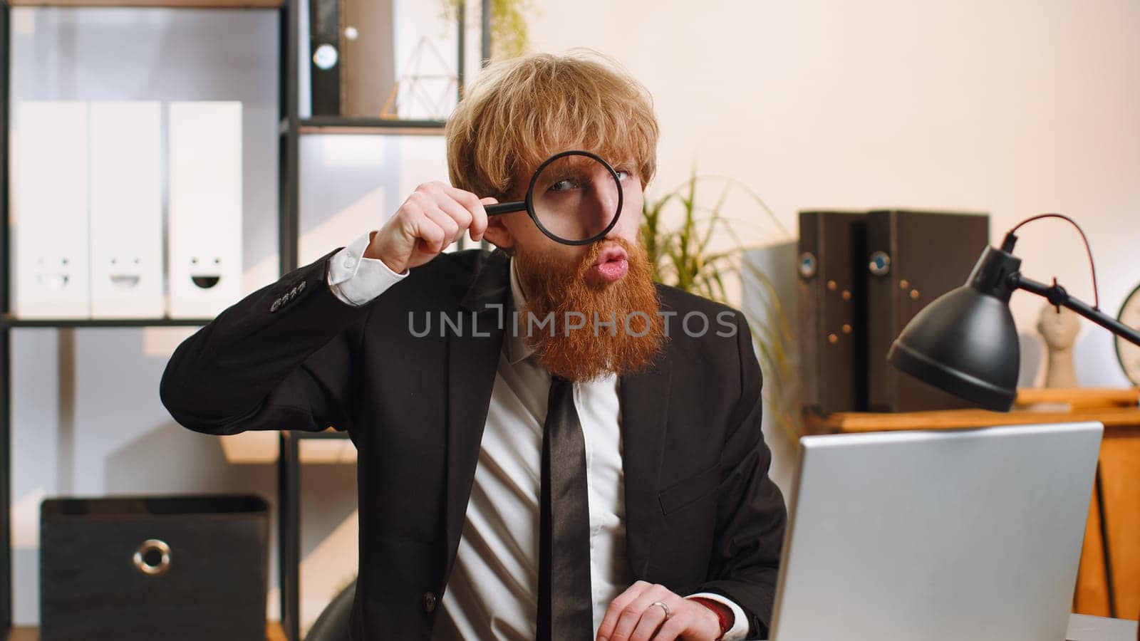 Investigator researcher scientist businessman working at home office holding magnifying glass near face looking into camera with big zoomed funny eye, searching, analysing. Professional freelancer man