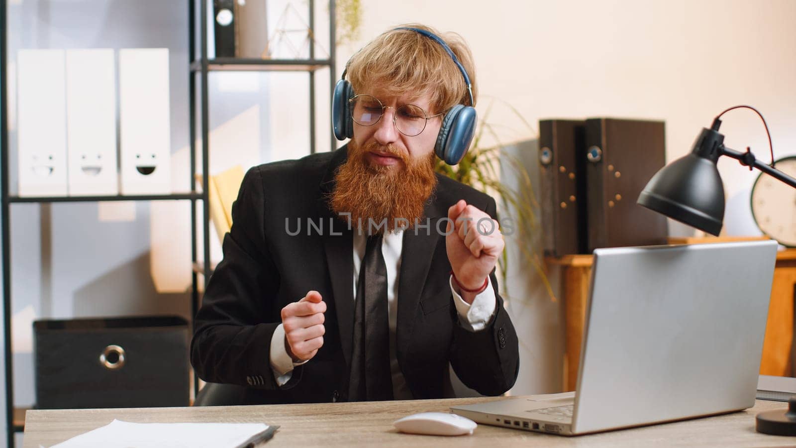 Happy relaxed overjoyed young businessman working on laptop computer at home office wearing headphones listening favorite energetic disco music on smartphone. Freelancer man relaxing, taking a break