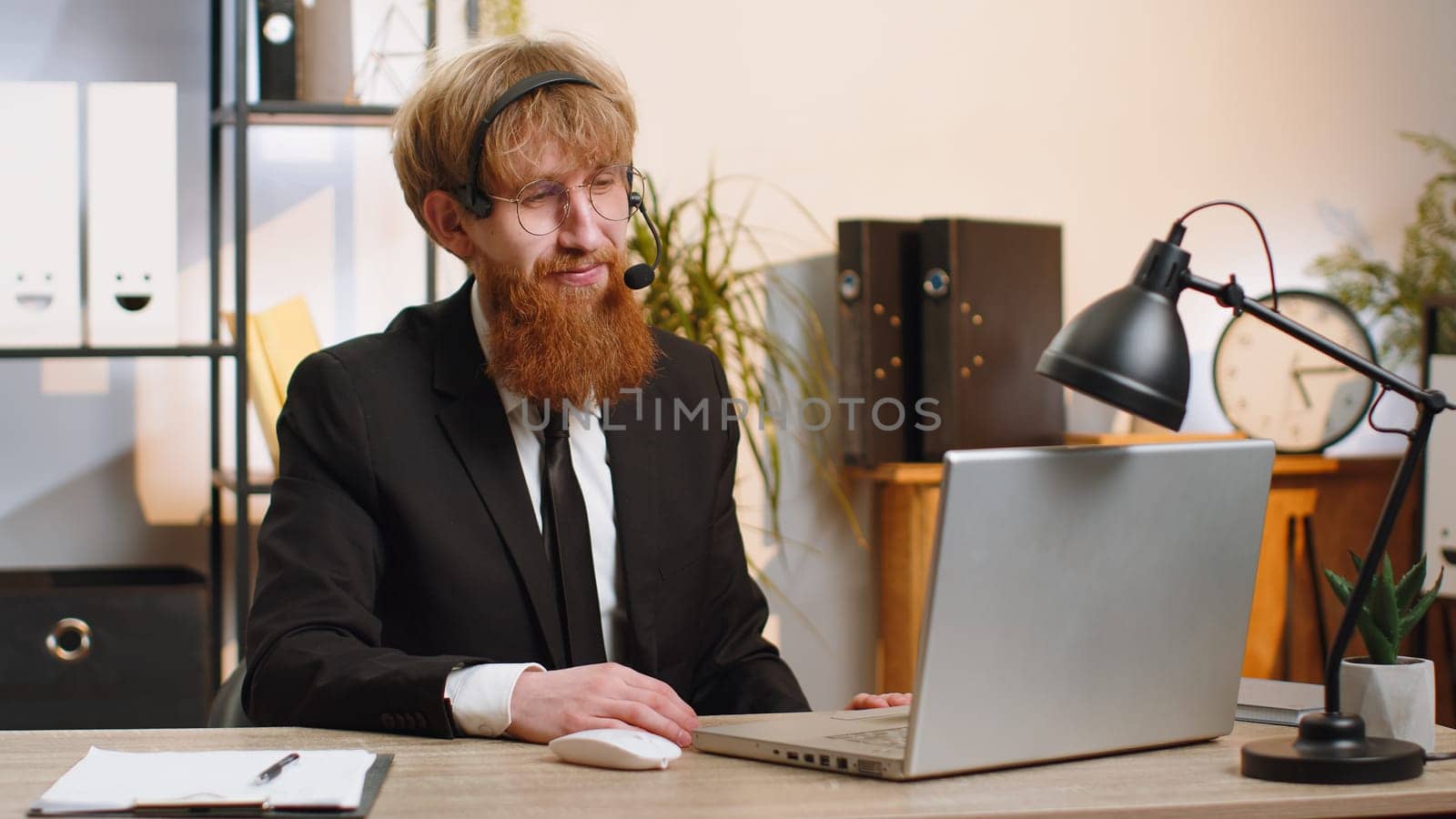 Young business man working on laptop wearing headset, freelance worker call center or support service operator helpline, having pleasant talk with client or colleague communication support at office
