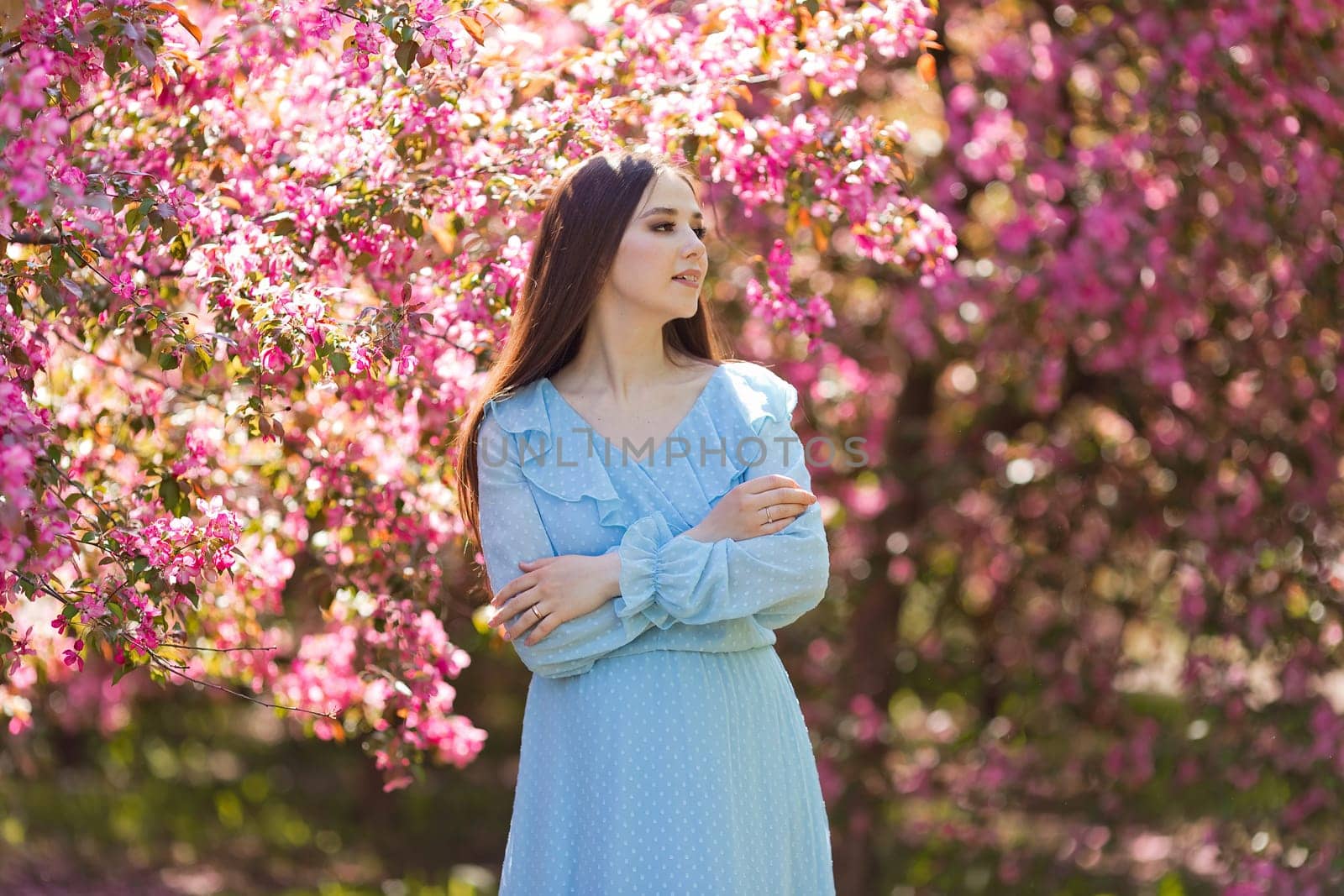 A pretty girl, brunette long hair, in light blue dress, standing in a pink blooming garden, looking away. Close up. Copy space