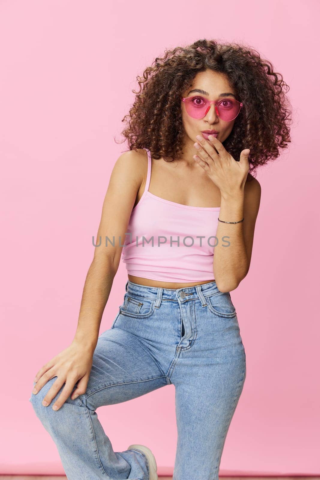 Happy woman afro curls hair dancing on a pink background in summer pink t-shirt jeans and glasses, summer vibe, copy space by SHOTPRIME