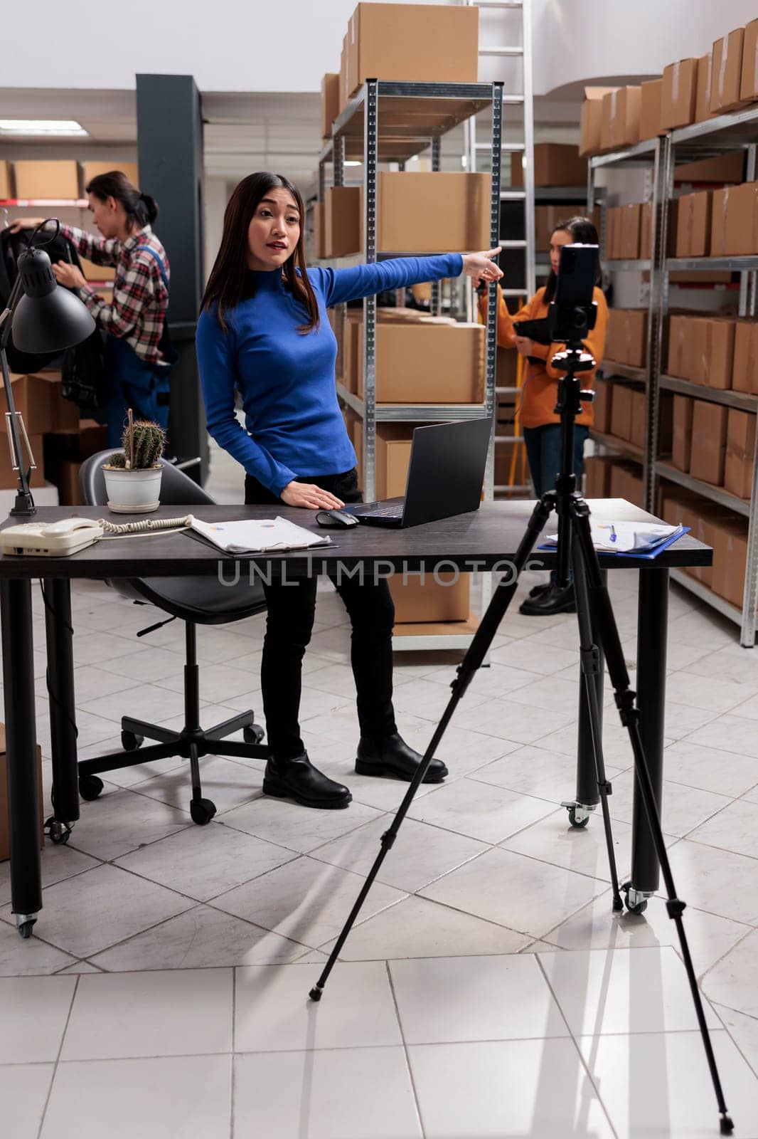 Asian woman recording employee testimonial video in industrial warehouse. Storehouse manager filming delivery operator and order picker coworkers team duties on mobile phone camera