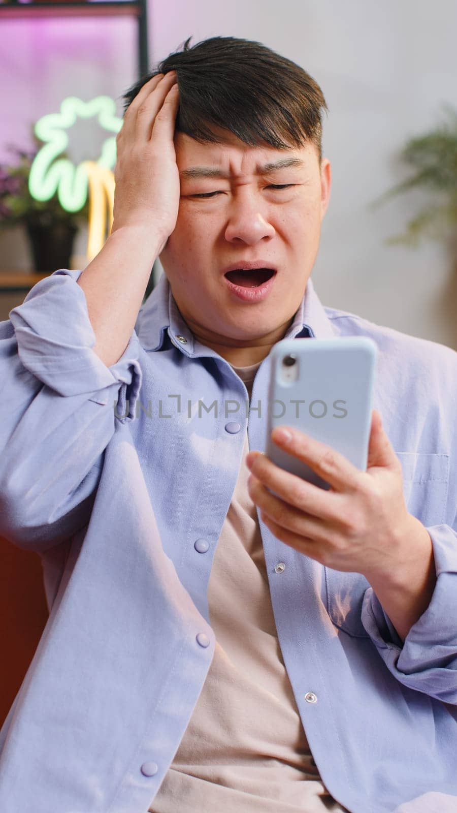 Sad asian displeased man use smartphone typing browsing, loses becoming surprised sudden lottery results, bad news, fortune loss, fail, deadline, virus. Chinese guy at home room. Vertical shot