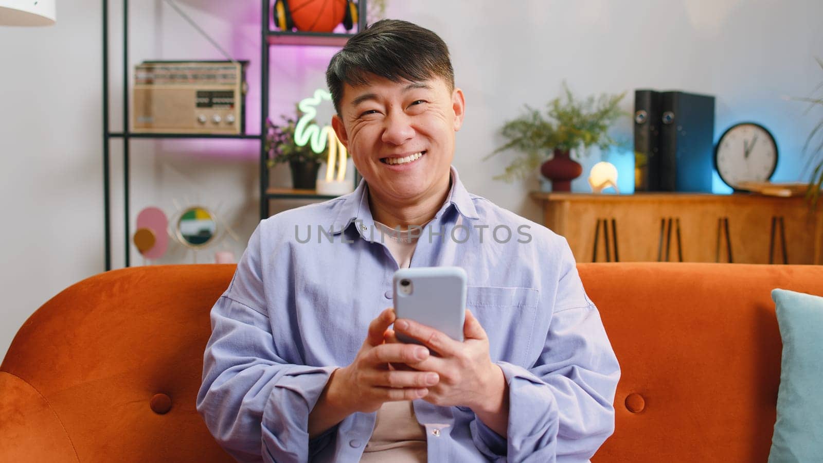 Asian man sitting on sofa uses mobile phone smiles at home living room apartment. Chinese adult guy texting share messages content on smartphone social media applications online, watching relax movie