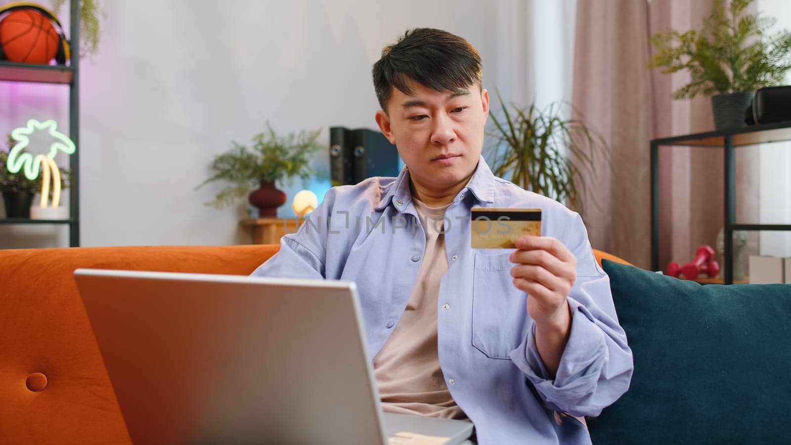 Asian man using credit bank card and laptop computer while transferring money, purchases online shopping, order food delivery at home apartment. Adult guy in day living room sitting on couch indoors